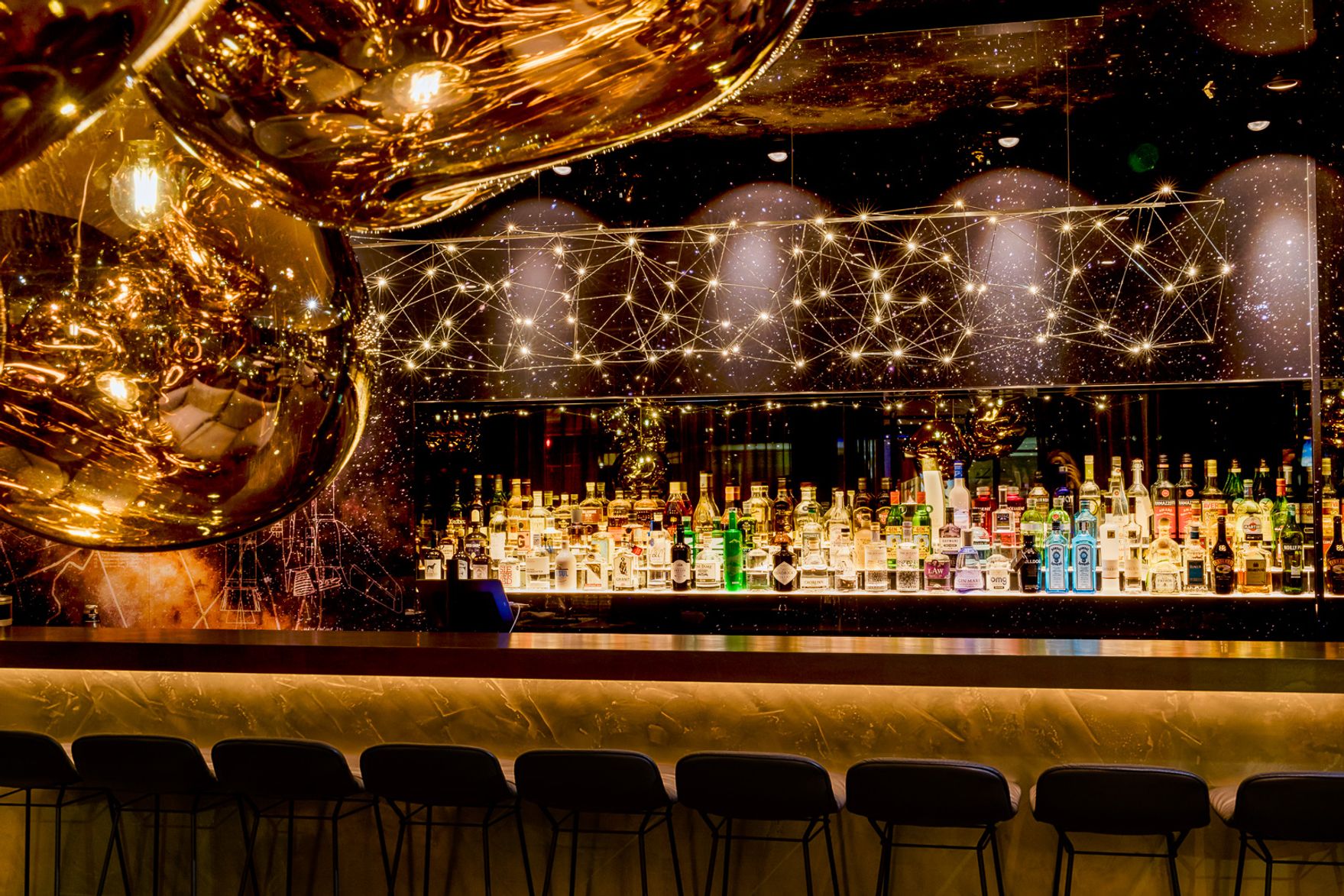Elegante Hotelbar mit stilvoll beleuchtetem Tresen, modernen goldfarbenen Pendelleuchten und einer großen Auswahl an Spirituosen im Hintergrund. Sitzplätze an der Bar laden zum entspannten Verweilen ein.