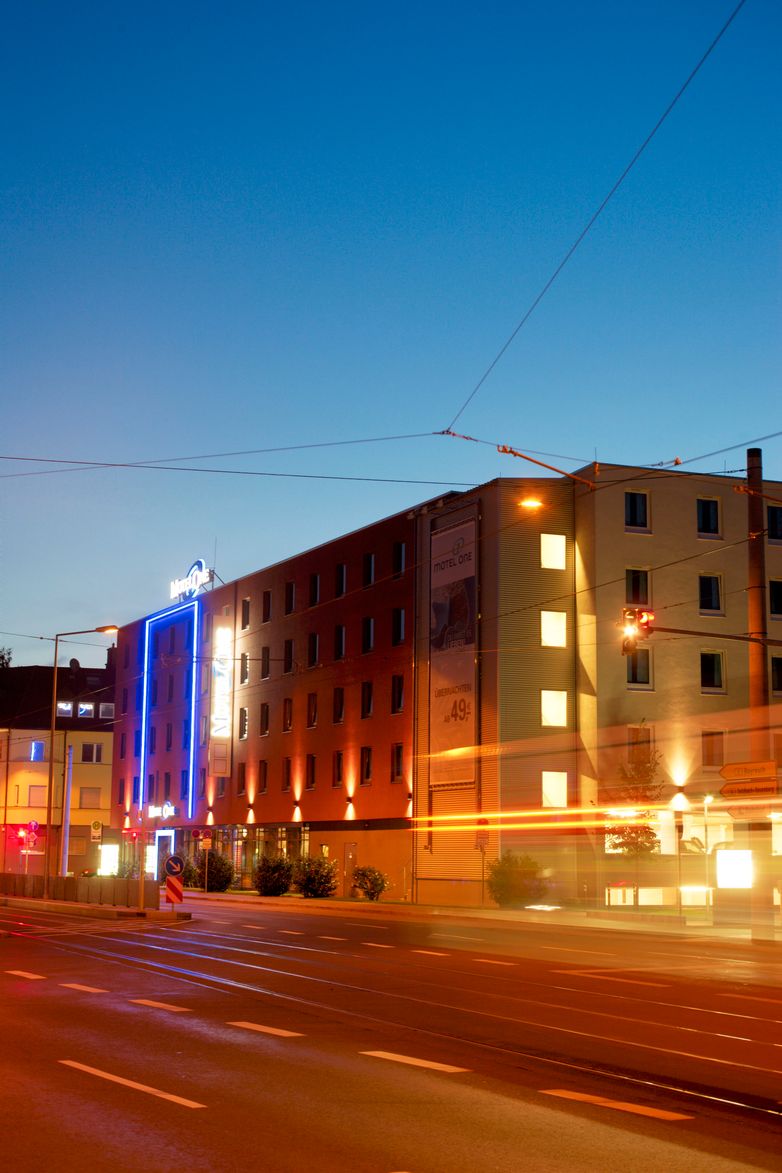 Fassade eines Hotels bei Abenddämmerung, beleuchtet mit warmem Licht und blauer Neonumrahmung, an einer ruhigen Straßenkreuzung in der Stadt.