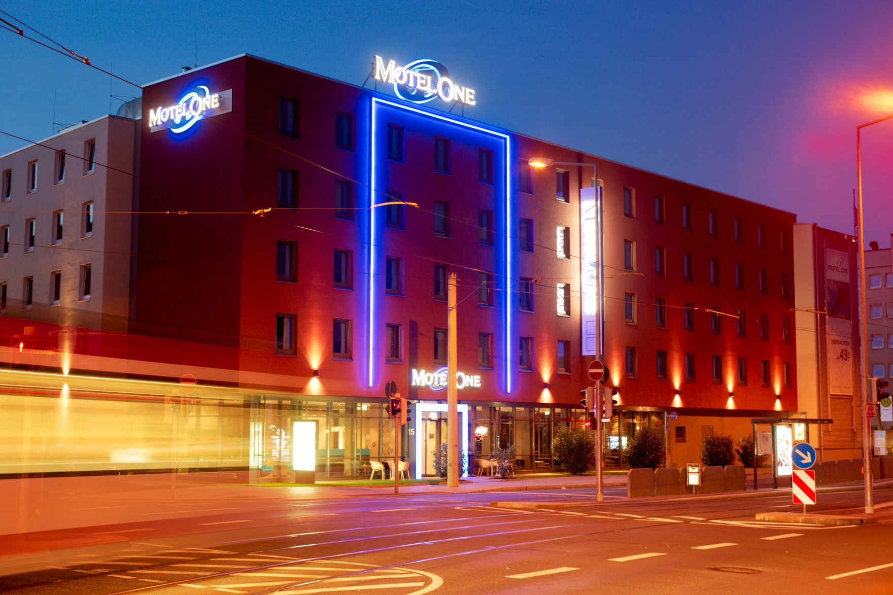 Außenansicht des Motel One Hotels bei Abenddämmerung mit beleuchtetem Hotellogo und hell erleuchtetem Eingangsbereich an einer städtischen Kreuzung.
