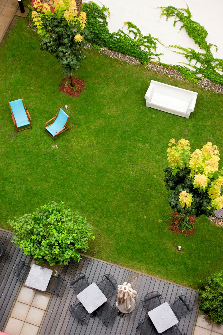 Blick von oben auf einen gepflegten Garten mit grüner Wiese, zwei Liegestühlen, einer weißen Gartenbank und Sitzbereichen auf einer Holzterrasse, umgeben von blühenden Büschen und Kletterpflanzen an einer weißen Wand.