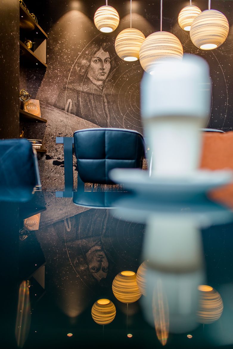 Gemütliche Lounge mit dunklen Sesseln, warmen Pendelleuchten und einer Wandkunst, die ein historisches Portrait und astronomische Motive zeigt. Spiegelnde Tischoberfläche verstärkt das stimmungsvolle, entspannte Ambiente.
