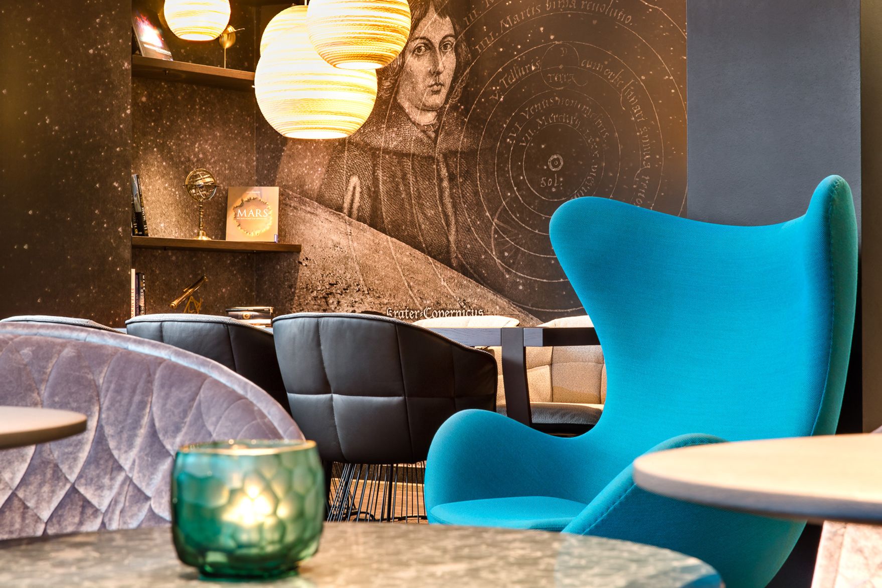 Moderne Lounge mit gemütlichen Sesseln, darunter ein auffälliger blauer Designstuhl, warmes Licht von hängenden Lampen und eine dekorative Wandgestaltung mit historischem Astronomie-Motiv für eine entspannte, stilvolle Atmosphäre.