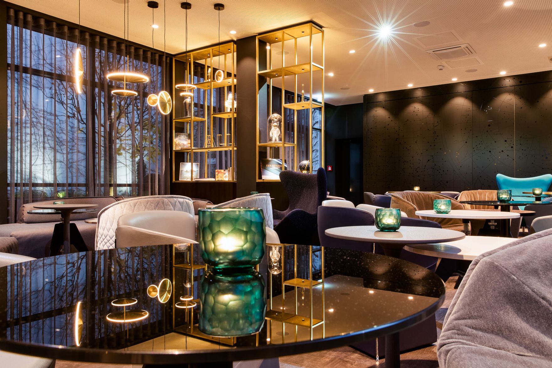 Moderner Hotel-Lounge-Bereich mit stilvollen Sitzmöbeln, stimmungsvoller Beleuchtung und dekorativen grünen Kerzenhaltern auf schwarzen Tischen, umgeben von großen Fenstern mit Blick auf Abenddämmerung und ruhiger Atmosphäre.