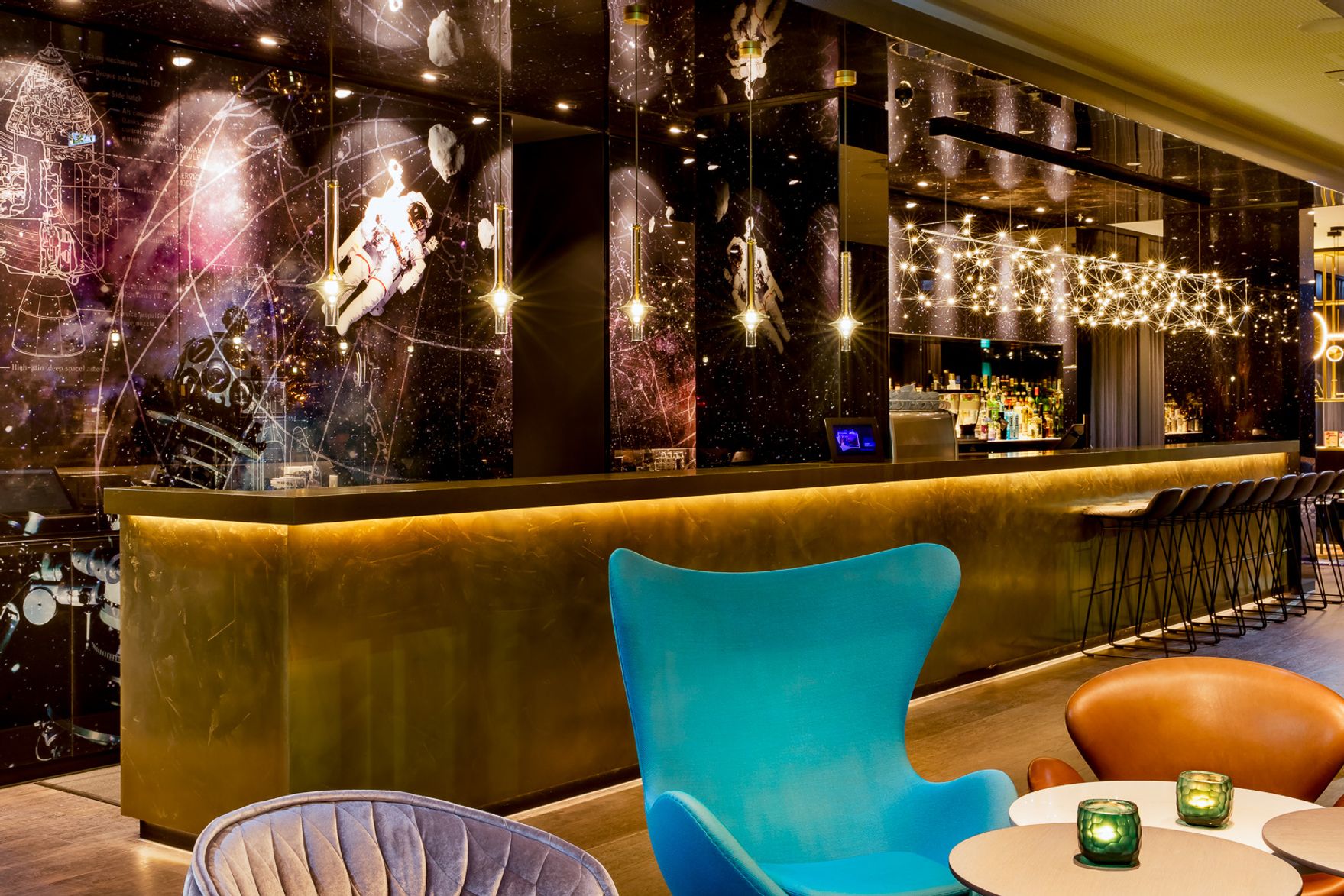 Hotelbar mit goldfarbenem Tresen, futuristischer Wanddekoration mit Weltraummotiven und gemütlichen Sitzgelegenheiten in verschiedenen Farben, einladend für entspannte Abende und gesellige Treffen.