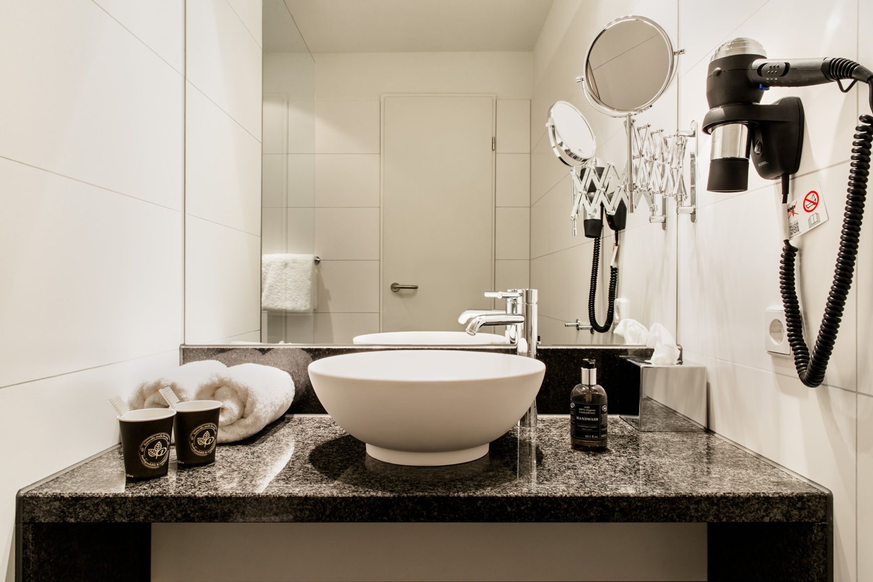 Moderne Hotelbadezimmer mit ovalem Waschbecken auf Granit, Wandspiegel, Wandföhn, vergrößerndem Kosmetikspiegel und frischen Handtüchern für komfortable Nutzung.