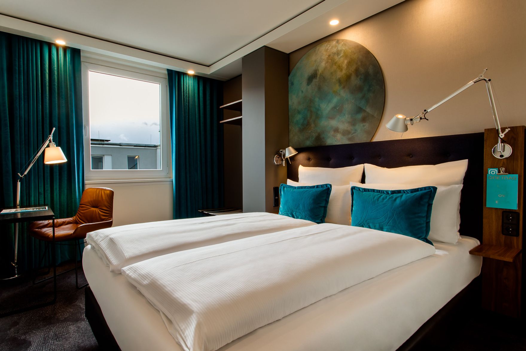 Gemütliches Hotelzimmer mit großem Doppelbett, weißen Bettdecken, blauen Akzentkissen, modernem Leselicht und einem großen Fenster mit dunkelgrünen Vorhängen, das sanftes Tageslicht hereinlässt.