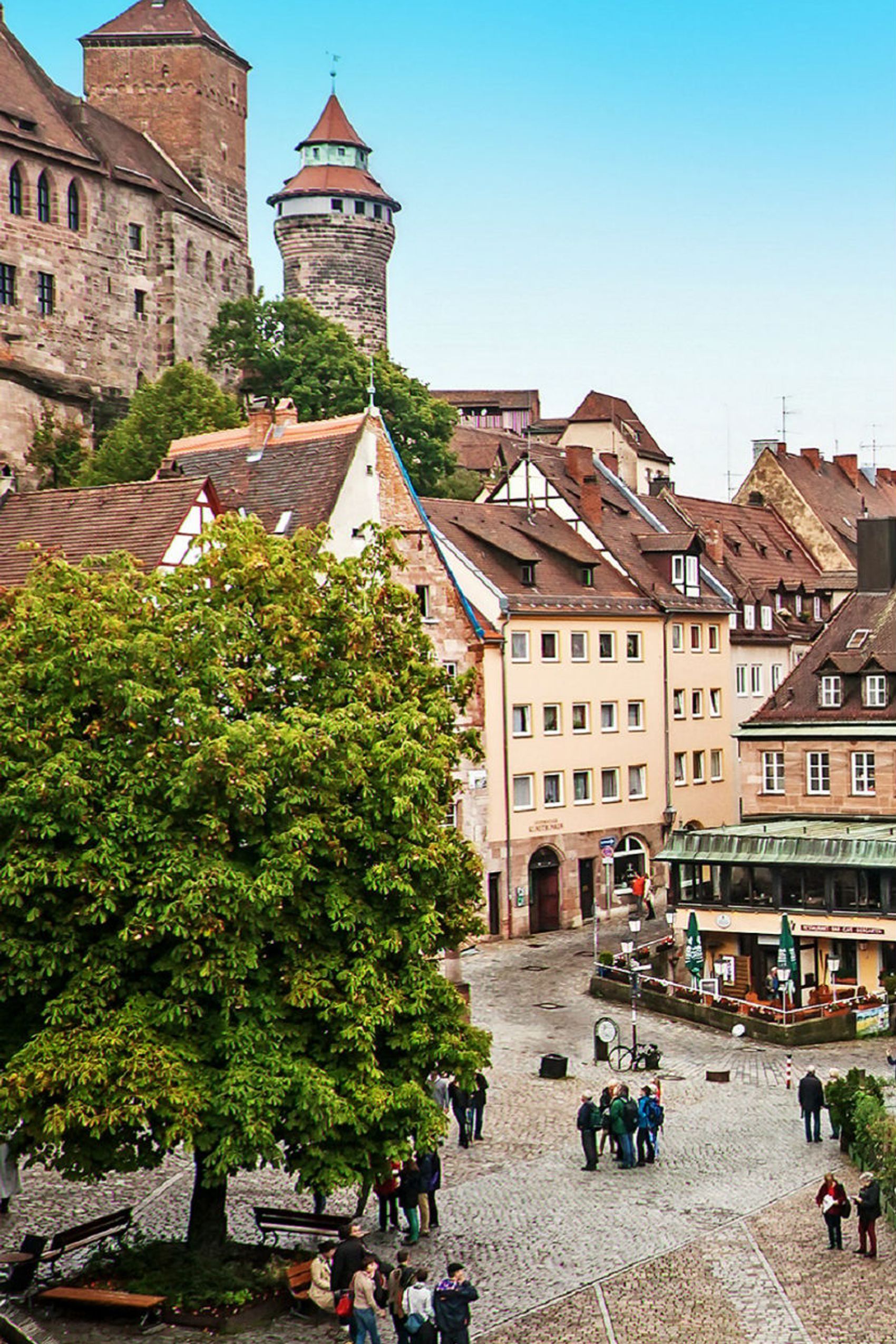 Blick auf eine historische Altstadt mit Kopfsteinpflaster, Fachwerkhäusern und mittelalterlichen Türmen unter klarem, blauem Himmel. Spaziergänger sind auf dem Platz unterwegs, der von Bäumen und gemütlichen Cafés gesäumt ist.