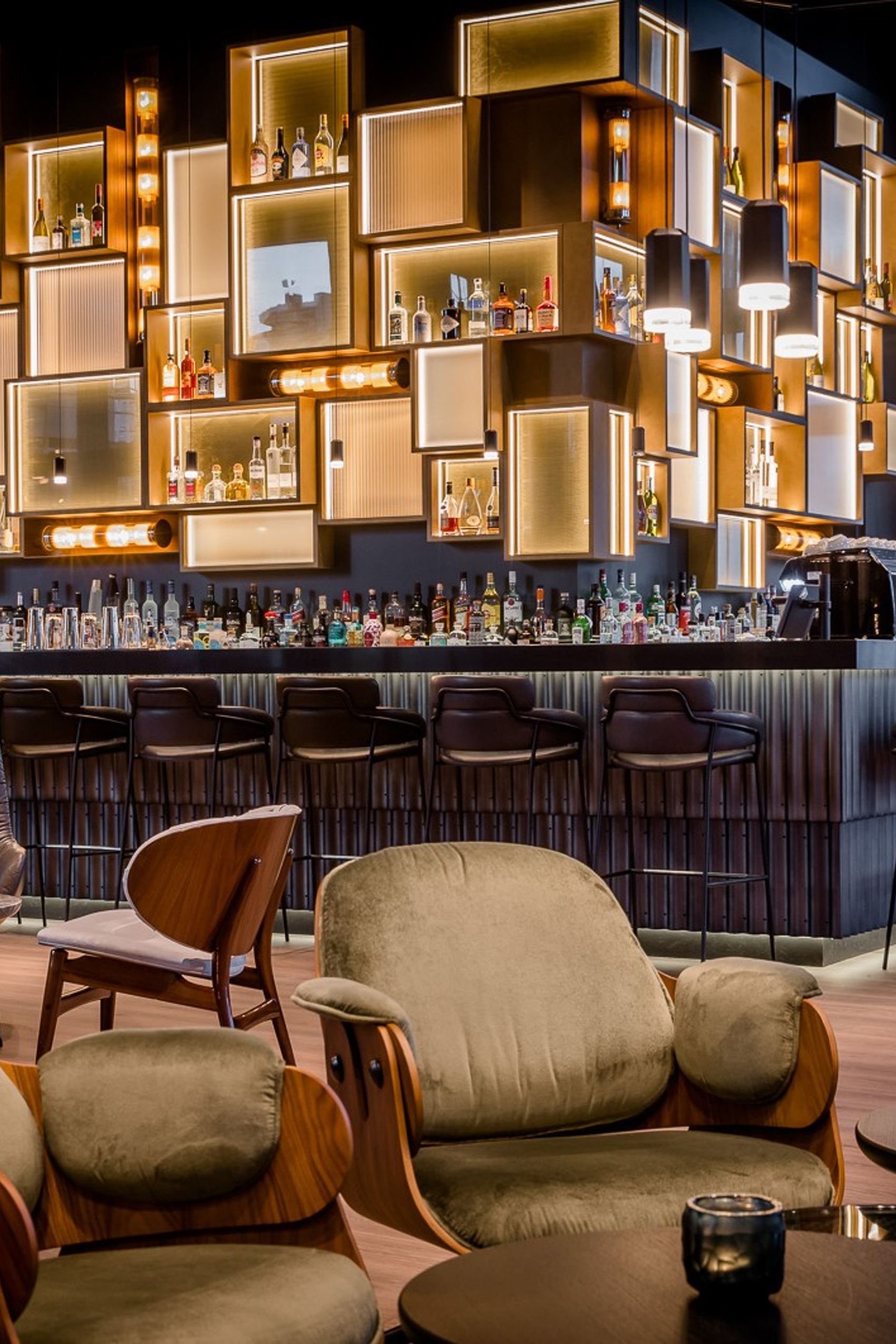 Hotelbar mit stilvollem, beleuchtetem Regal aus geometrischen Elementen, bequemen Sesseln und Barhockern, die eine einladende Atmosphäre für entspannte Abende schafft.