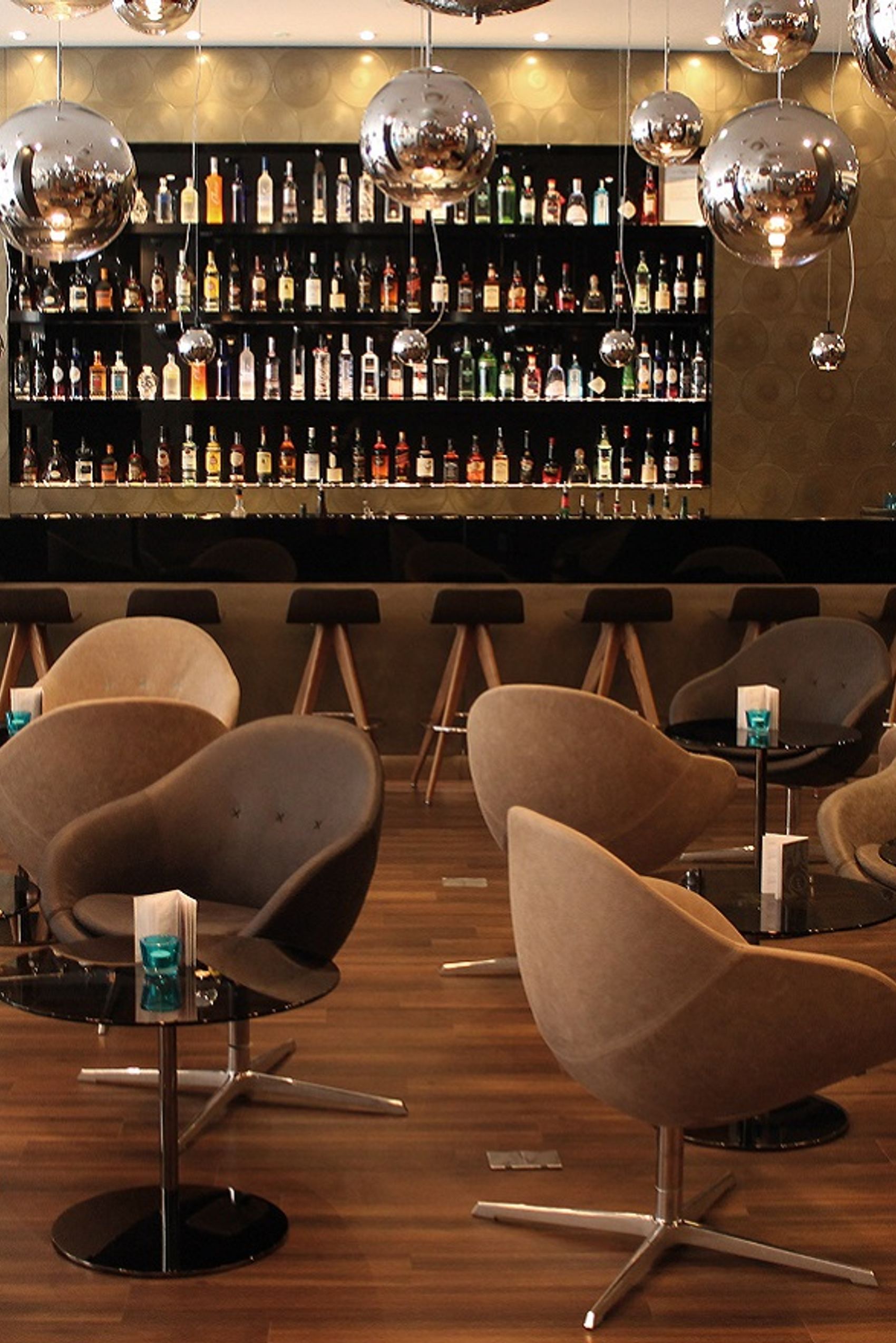 Hotelbar mit stilvollen, bequemen Sesseln und dunkler Theke. Große Auswahl an Spirituosen hinter der Bar, atmosphärische Beleuchtung durch glänzende, runde Pendelleuchten.