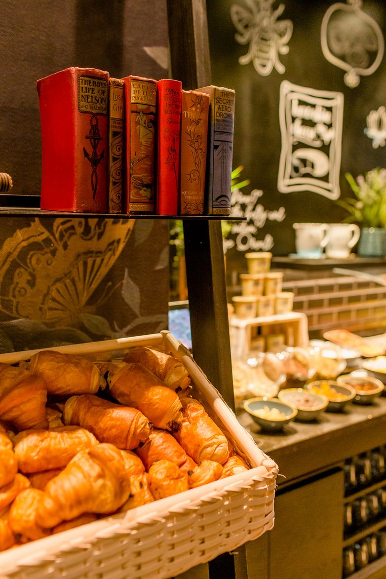 Korb mit frischen Croissants vor einem Frühstücksbuffet, im Hintergrund dekorative Bücher und eine Tafelwand mit Zeichnungen, warme und einladende Atmosphäre.