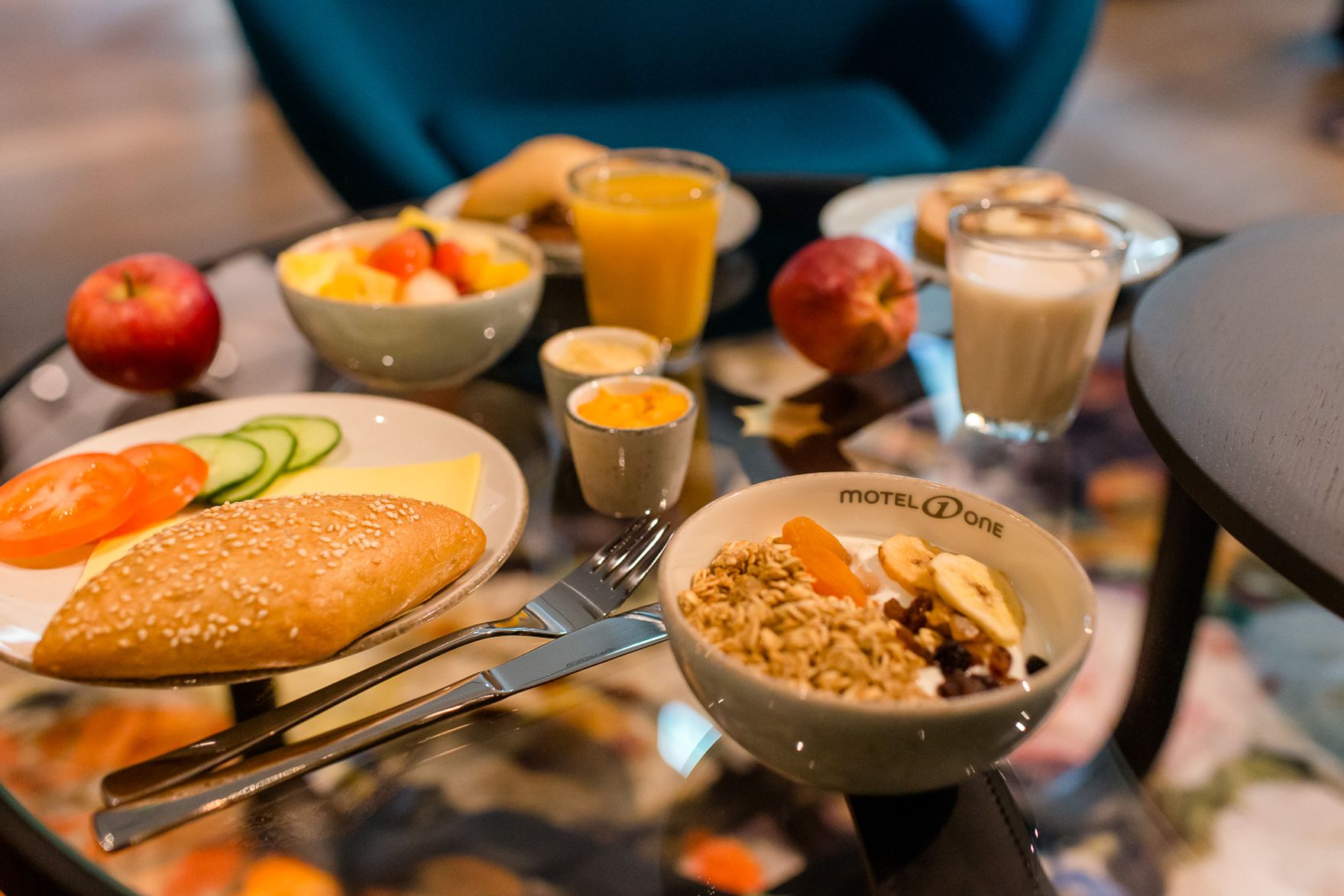 Frühstückstisch mit frischem Brötchen, Müsli mit Obst, frischem Obst, Gemüse, zwei Kaffeegetränken und Orangensaft, präsentiert auf einem Glas-Tisch.