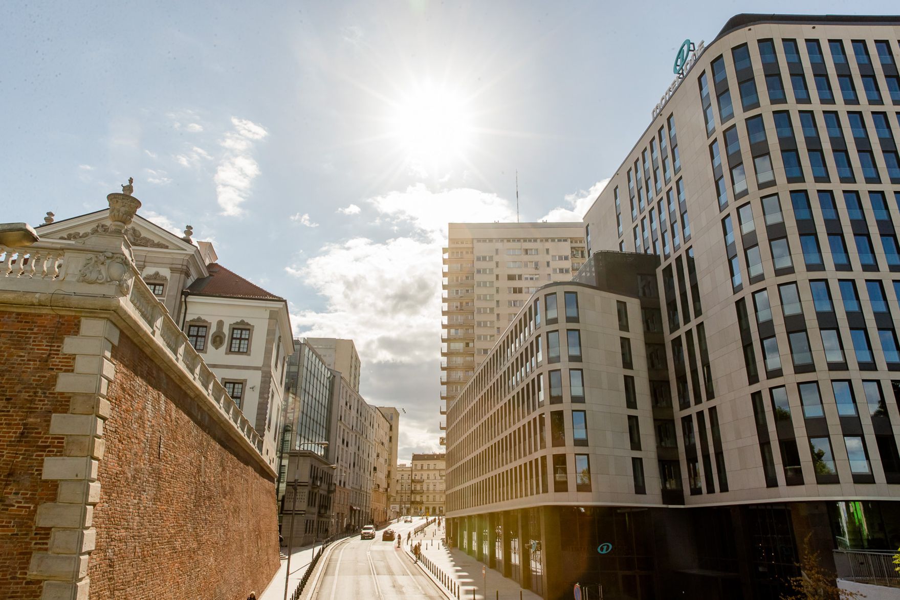 Hotel Warschau Motel One sonniger Blick auf eine moderne Straßenfront mit hohen, gläsernen Hotelgebäuden und historischen Mauern, die Tradition und zeitgenössische Architektur verbinden.