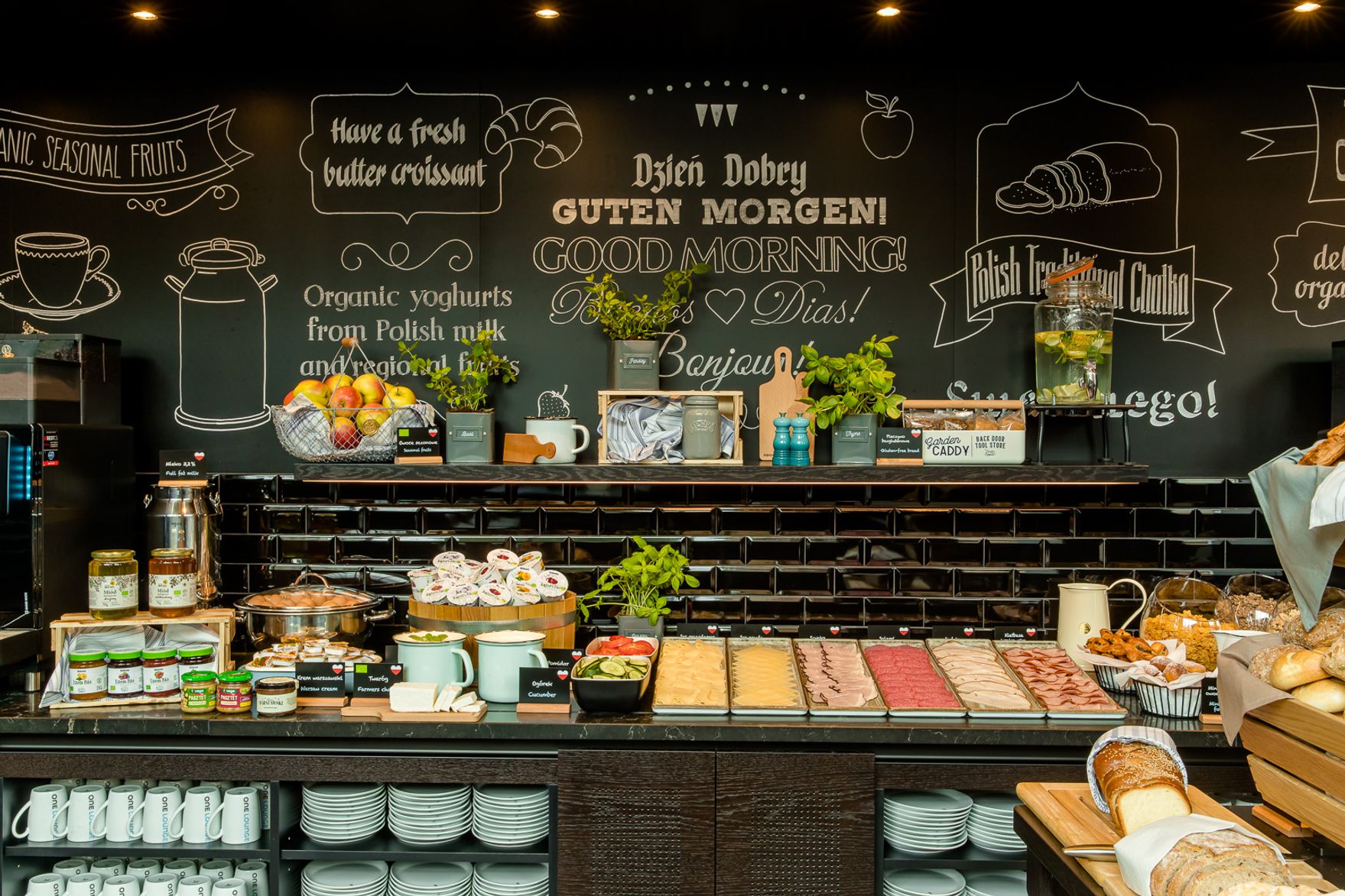 Frühstücksbuffet mit frischem Obst, Croissants, Aufschnitt, Joghurts und Getränken vor schwarzer Wand mit guten Morgen Grüßen und Hinweisen zu polnischen Spezialitäten.