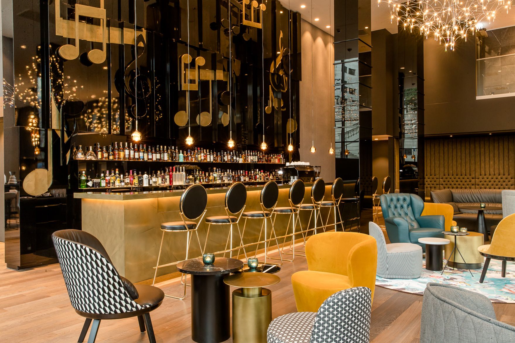 Moderne Hotelbar mit eleganter Theke, dunkelglänzenden Wänden und goldenen Noten-Dekorationen. Gemütliche Sitzgelegenheiten in warmen Farben laden zum Entspannen und geselligen Beisammensein ein. Stilvolle Beleuchtung schafft eine einladende Atmosphäre.