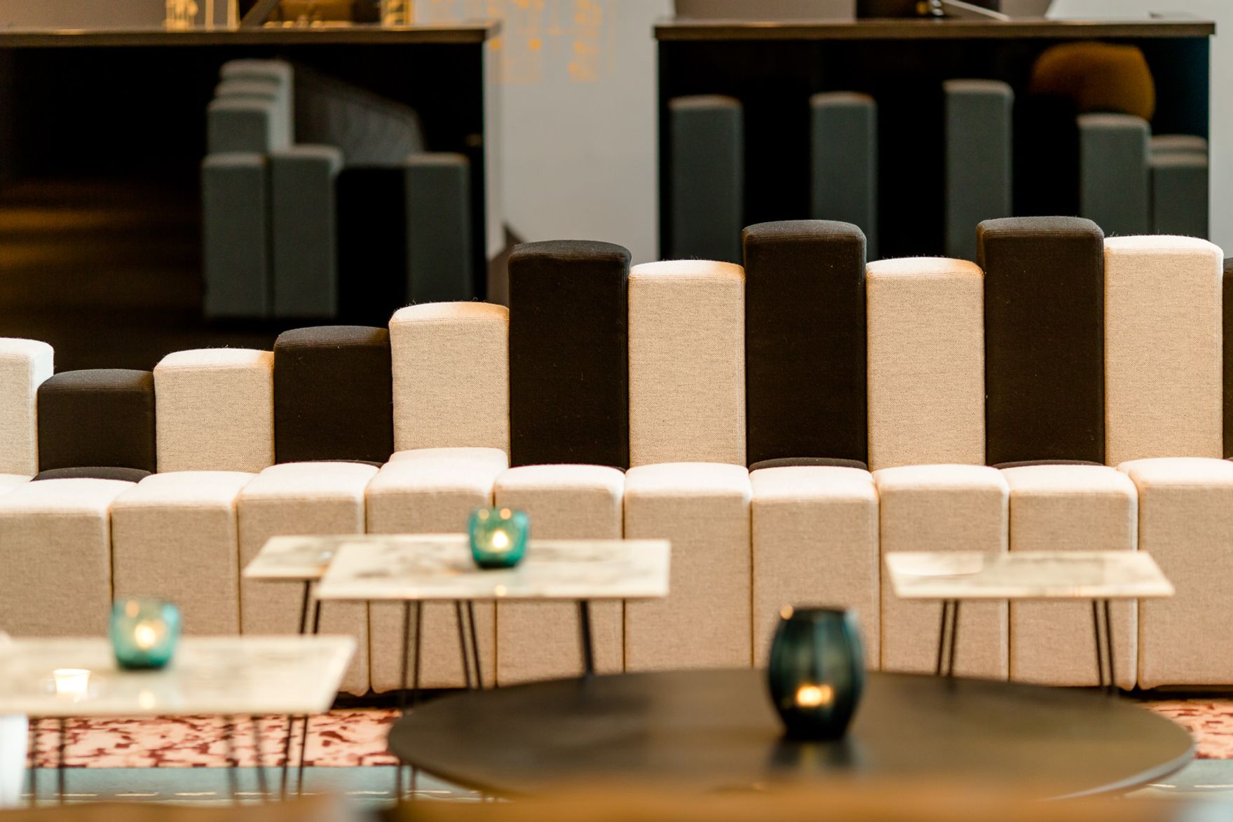 Moderne Lounge mit stilvoll gepolsterten Sitzbänken in abwechselnden Schwarz- und Beigetönen, umgeben von niedrigen Tischen und stimmungsvoller Beleuchtung durch kleine Kerzenhalter.