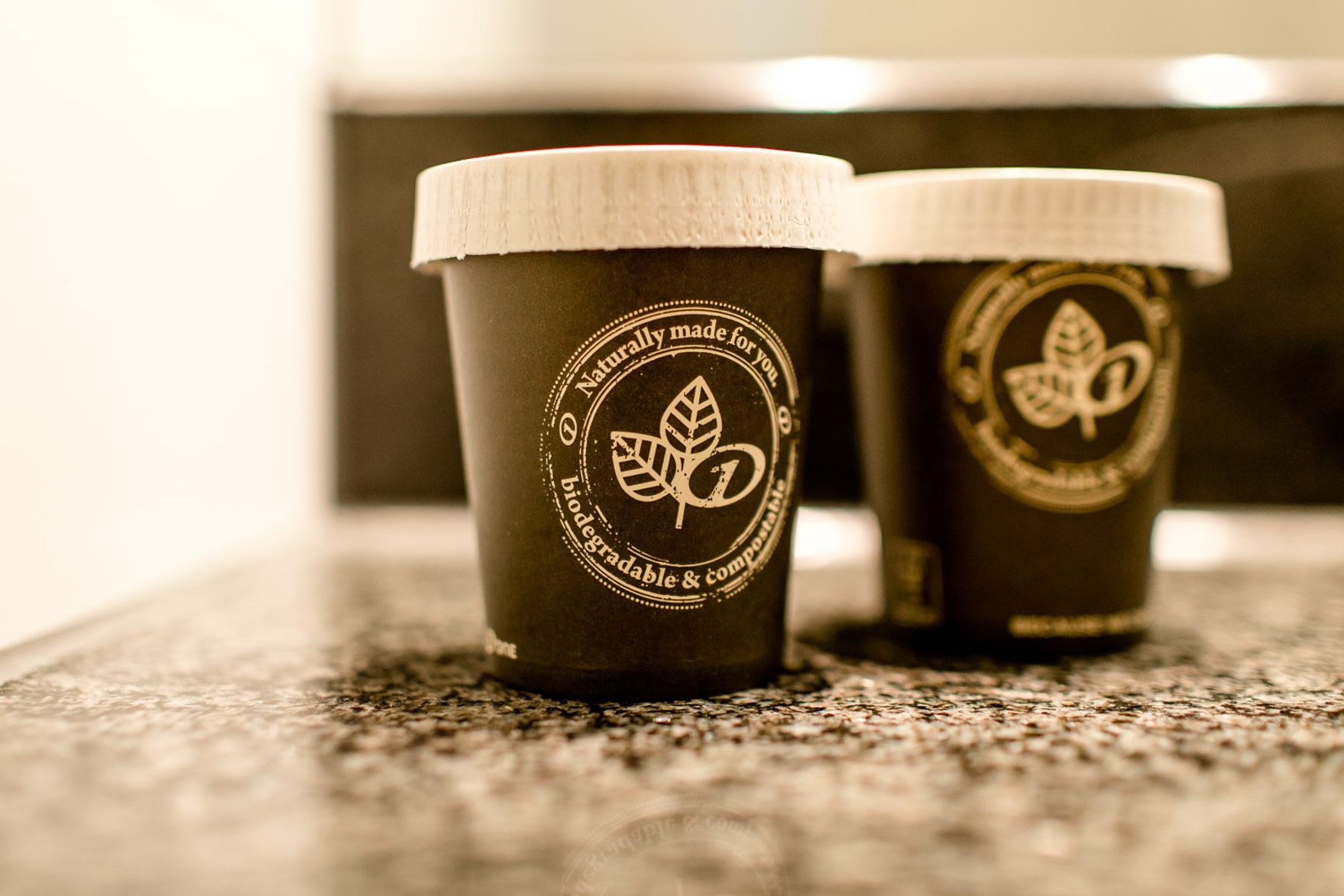 Zwei nachhaltige, biologisch abbaubare Kaffeebecher mit Deckel stehen auf einer Marmoroberfläche, symbolisieren umweltfreundlichen Kaffee-Genuss.