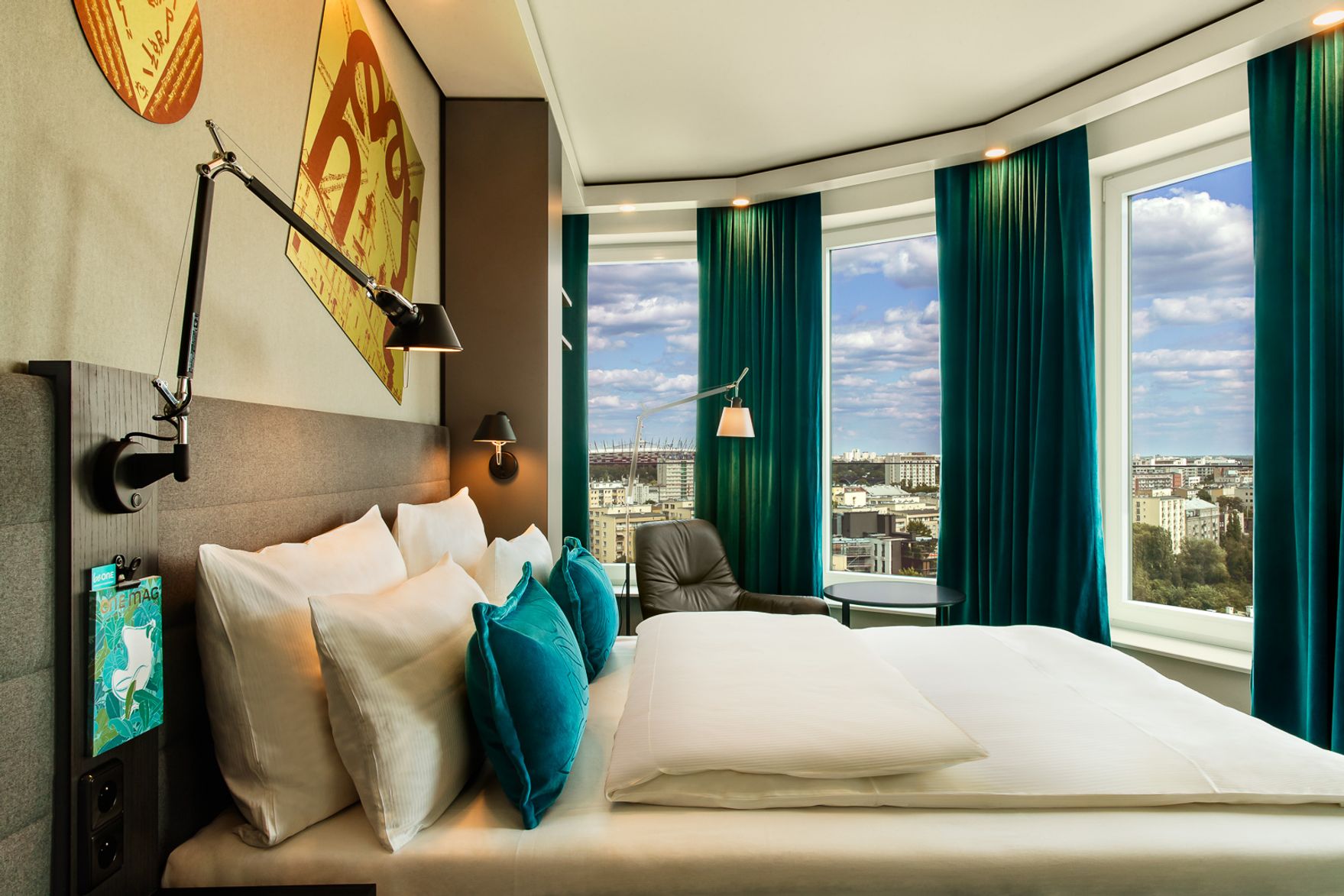 Modernes Hotelzimmer mit großem, gemütlichem Bett, mehreren Kissen und Panoramafenstern, die einen weiten Blick über die Stadt bieten. Dunkelgrüne Vorhänge rahmen das Tageslicht und schaffen eine einladende Atmosphäre zum Entspannen.