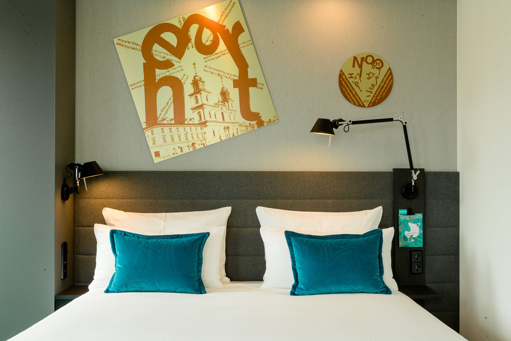 Modern eingerichtetes Hotelzimmer mit großem Doppelbett, zwei türkisfarbenen Kissen, Leselampen über dem Bett und stilvoller Wanddekoration in warmen Farbtönen für eine gemütliche Atmosphäre.