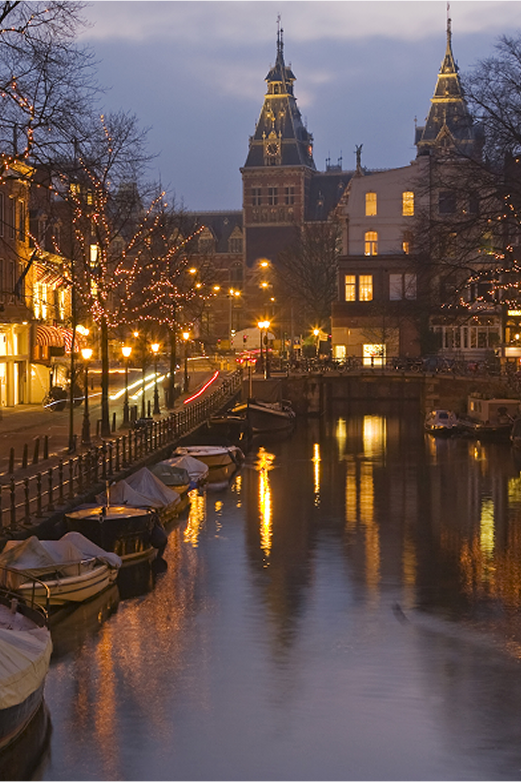 Abendliche Stadtansicht an einem ruhigen Kanal mit beleuchteten Gebäuden und kleinen Booten, die im Wasser liegen. Im Hintergrund sind historische Türme zu erkennen.