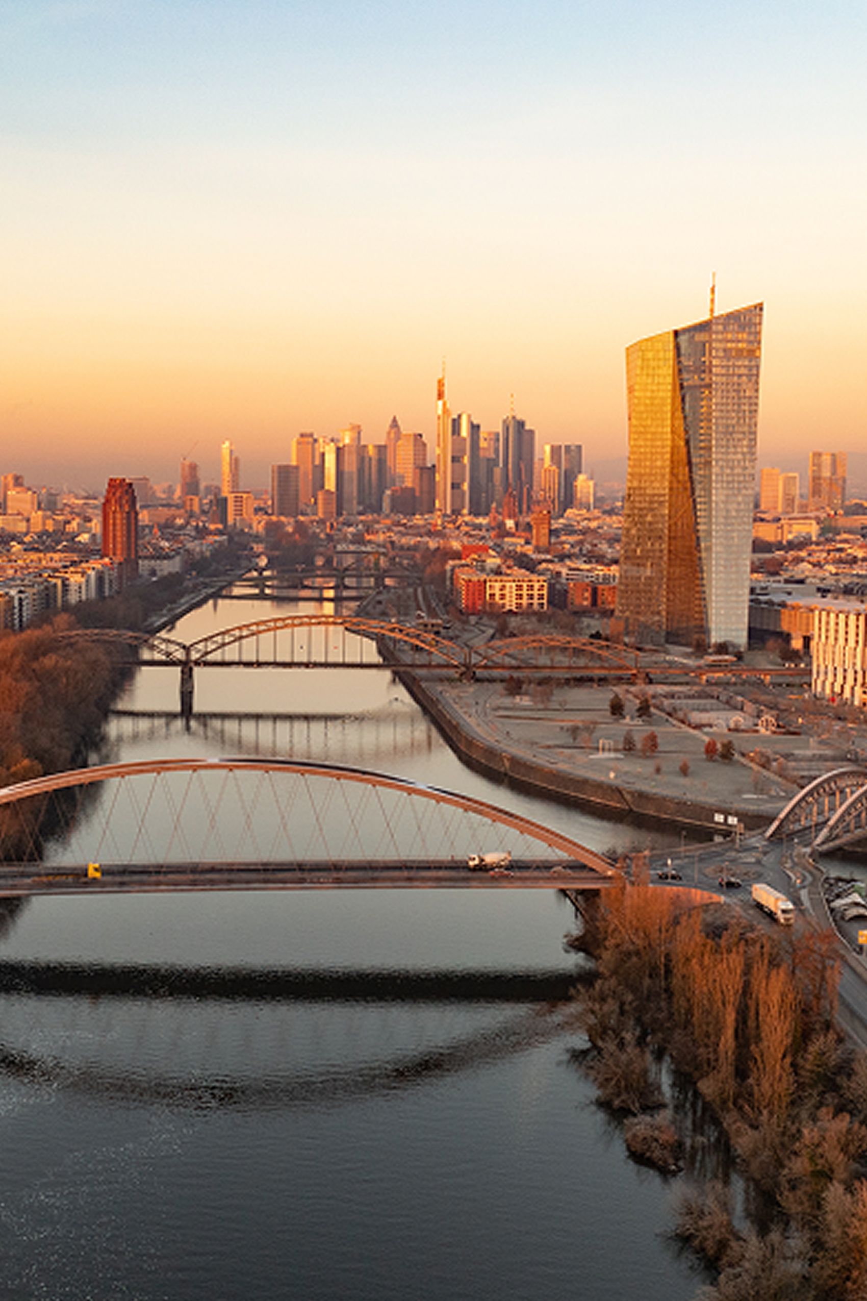 Blick auf einen Fluss mit mehreren Brücken, Hochhäusern im Hintergrund bei Sonnenuntergang, darunter ein markantes, goldglänzendes Hochhaus am Ufer, das eine urbane Skyline prägt.