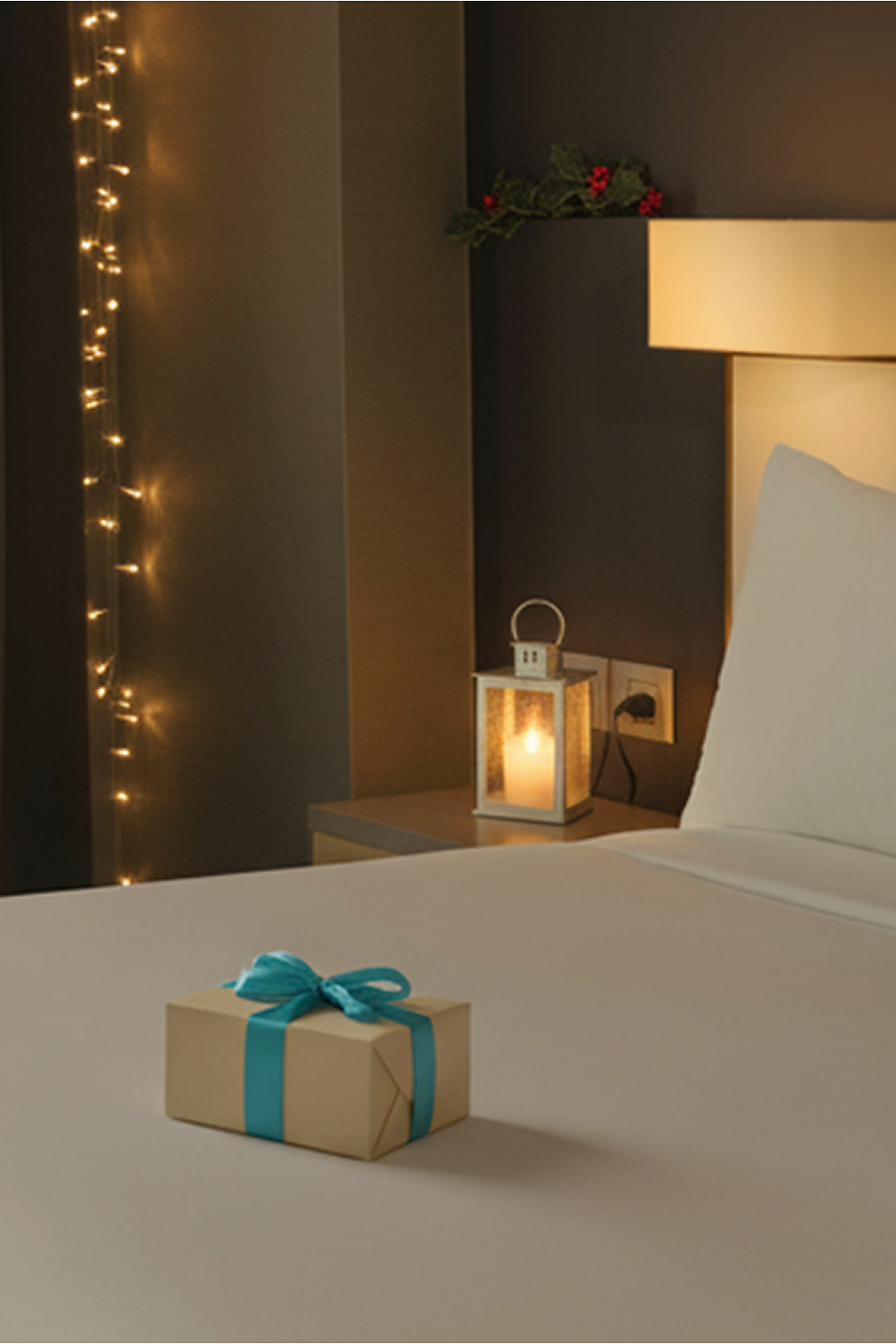 Ein gemütlich beleuchtetes Hotelzimmer mit weihnachtlicher Dekoration, einem warm leuchtenden Windlicht auf dem Nachttisch und einem liebevoll verpackten Geschenk auf dem Bett, das eine festliche Atmosphäre und Geborgenheit vermittelt.
