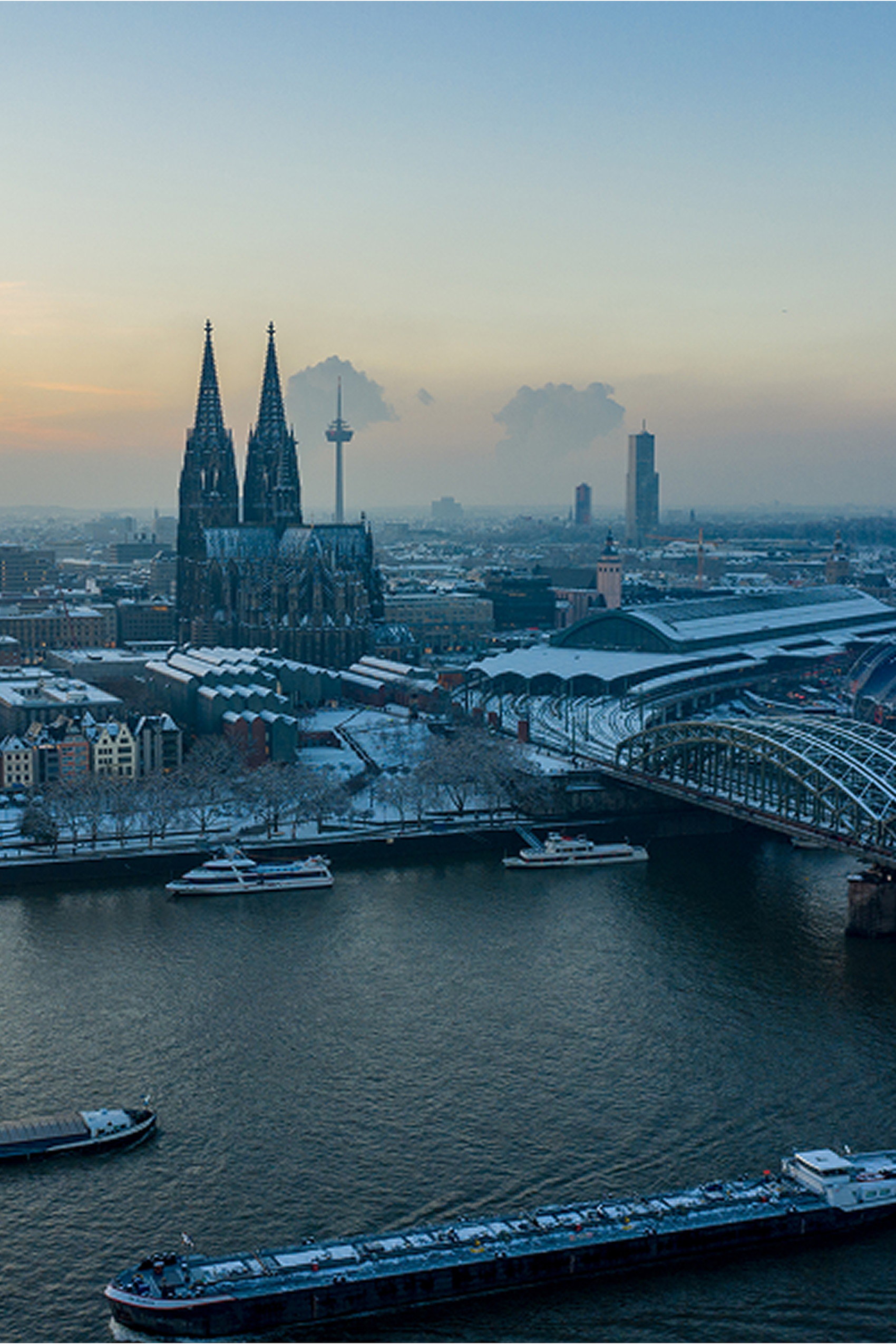 Blick über den Rhein auf den Kölner Dom bei Dämmerung, umgeben von Stadtgebäuden und Brücken.