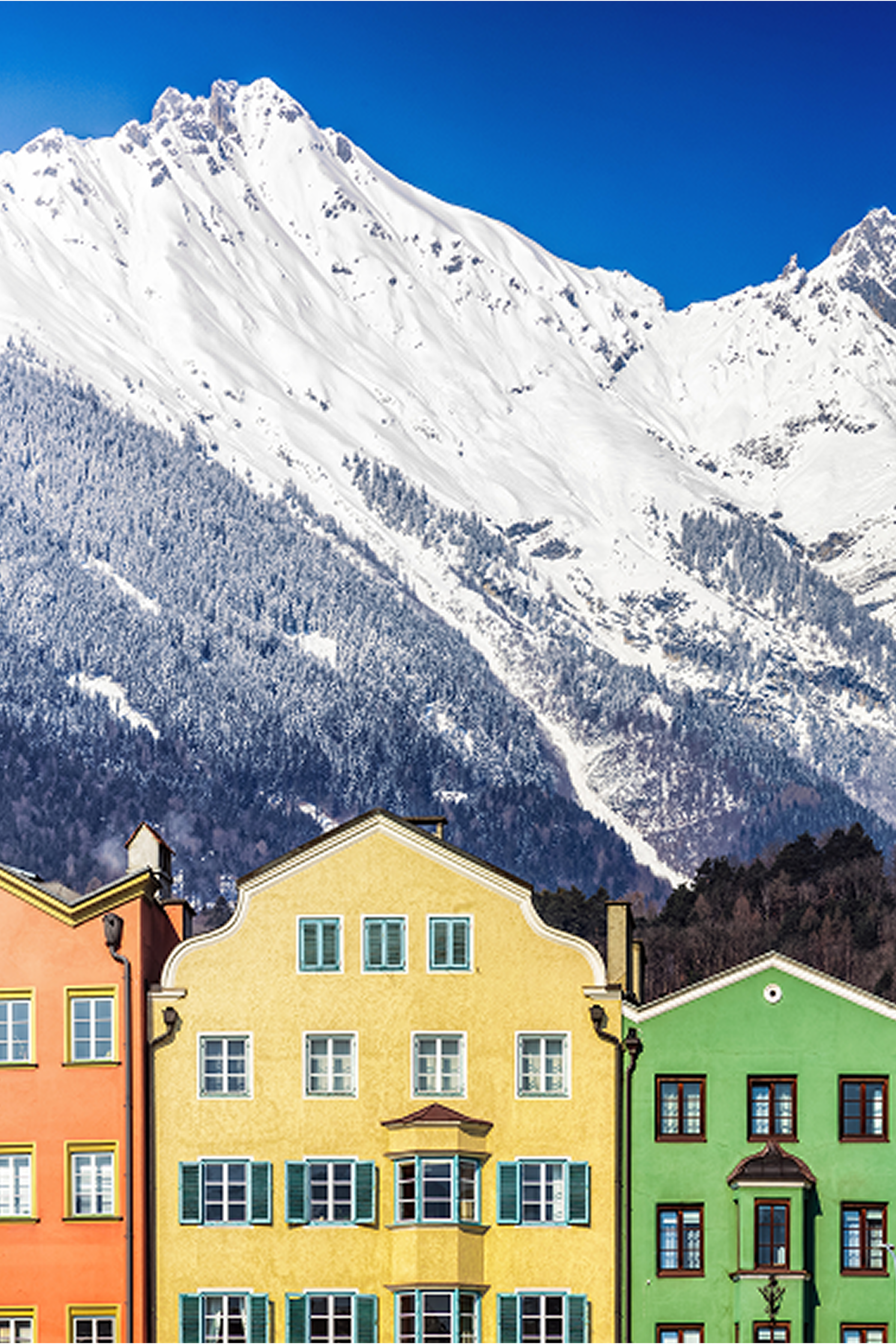 Bunte Häuserfront vor majestätischen, schneebedeckten Bergen unter klarem blauem Himmel, die eine malerische Alpenlandschaft mit charmantem Dorfambiente zeigt.