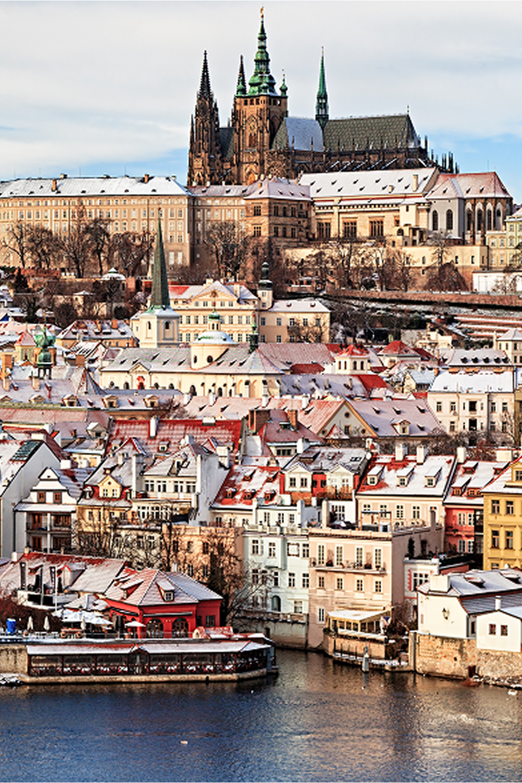 Vogelperspektive auf Prager Altstadt mit verschneiten Dächern, zahlreichen historischen Gebäuden und der markanten Prager Burg im Hintergrund, die die Skyline prägt. Flussfront mit Bootsanleger im Vordergrund. Winterliche Atmosphäre unter bewölktem Himmel.
