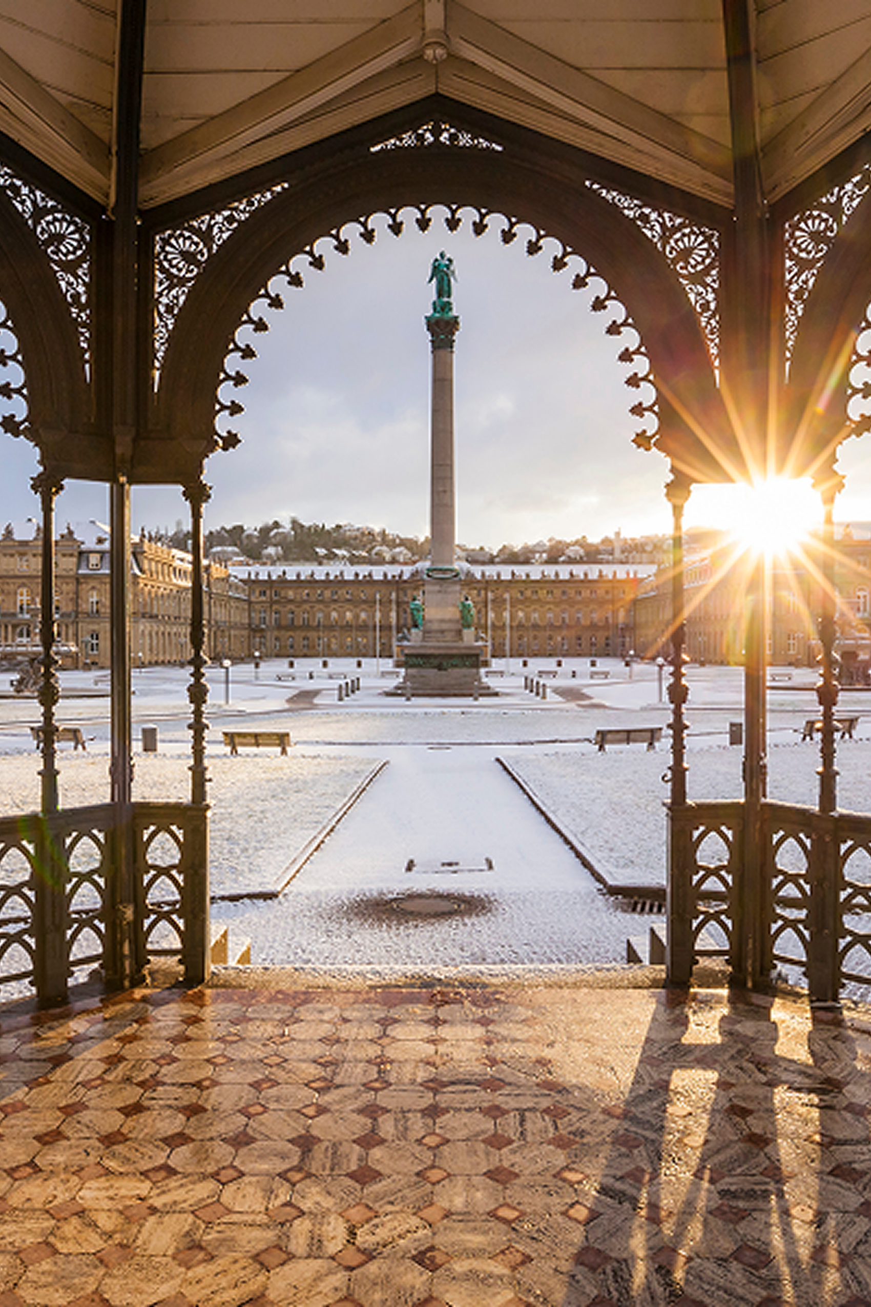 Blick von einem verzierten Pavillon auf einen verschneiten Platz mit einer hohen Säule und Statue, im Hintergrund historische Gebäude, die Morgensonne scheint seitlich und erzeugt warme Lichtstrahlen.