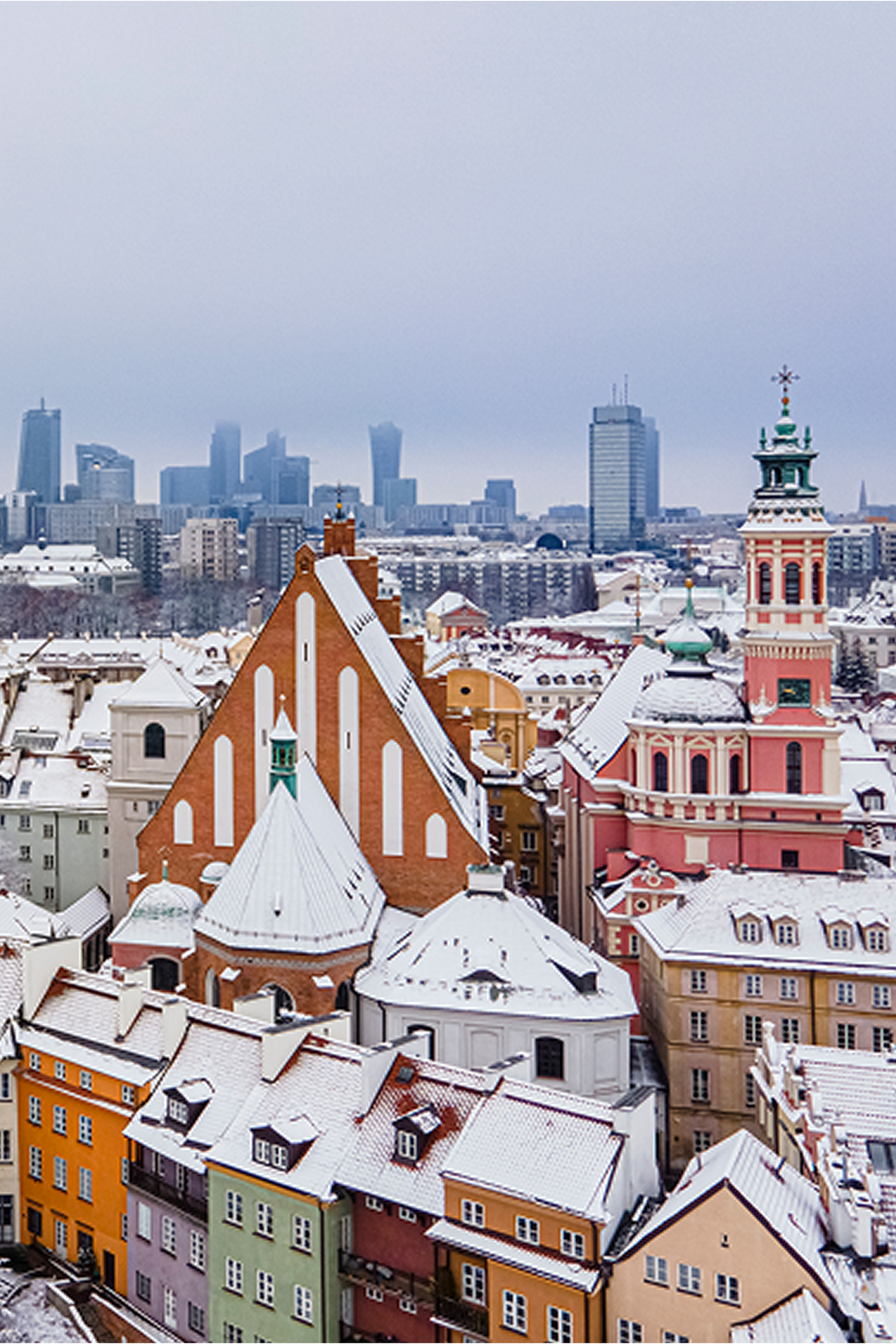 Blick auf eine verschneite Altstadt mit bunten Häusern und historischen Kirchen, im Hintergrund moderne Hochhäuser einer Großstadt unter einem bewölkten Himmel.