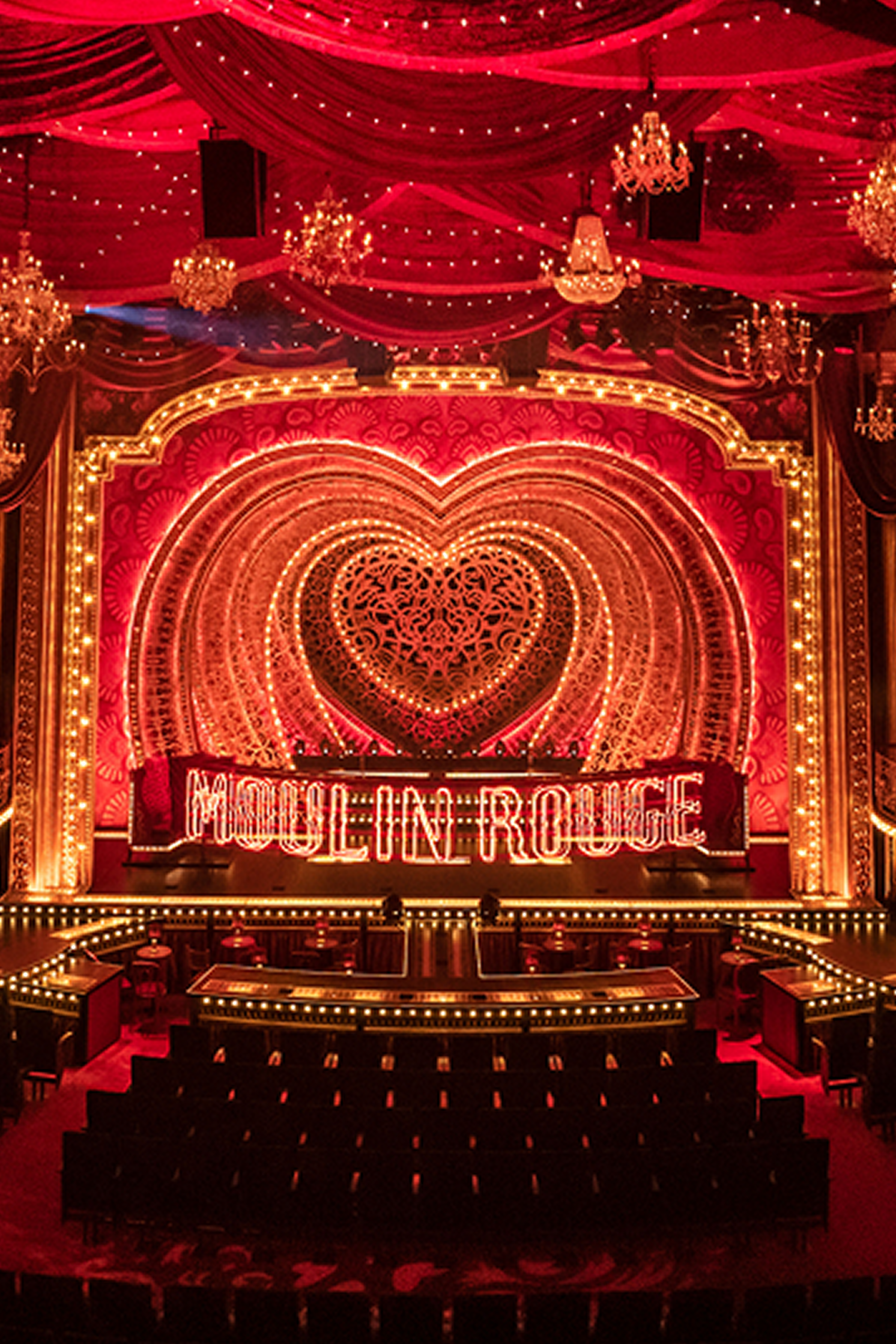 Blick auf die Bühne des Moulin Rouge mit leuchtenden, roten Herzmotiven, goldener Beleuchtung und rotem Samtvorhang, umgeben von funkelnden Kronleuchtern.