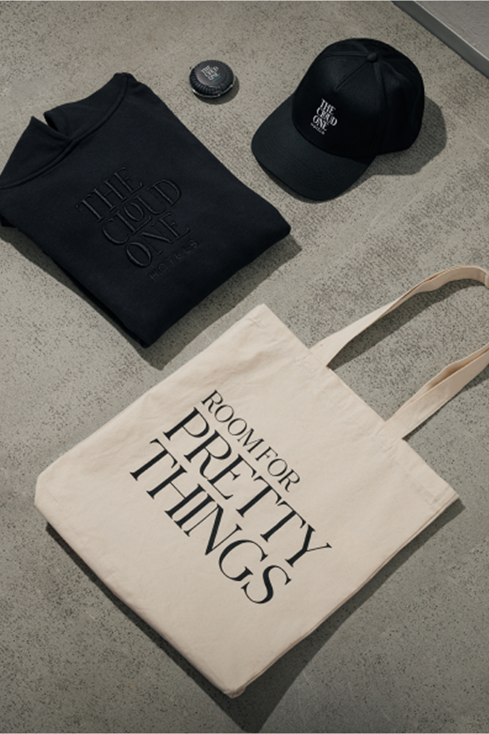 Schwarzes Sweatshirt und Cap mit der Aufschrift „THE COLD ONE“ sowie eine helle Stofftasche mit dem Schriftzug „ROOM FOR PRETTY THINGS“ liegen auf einem grauen Boden.
