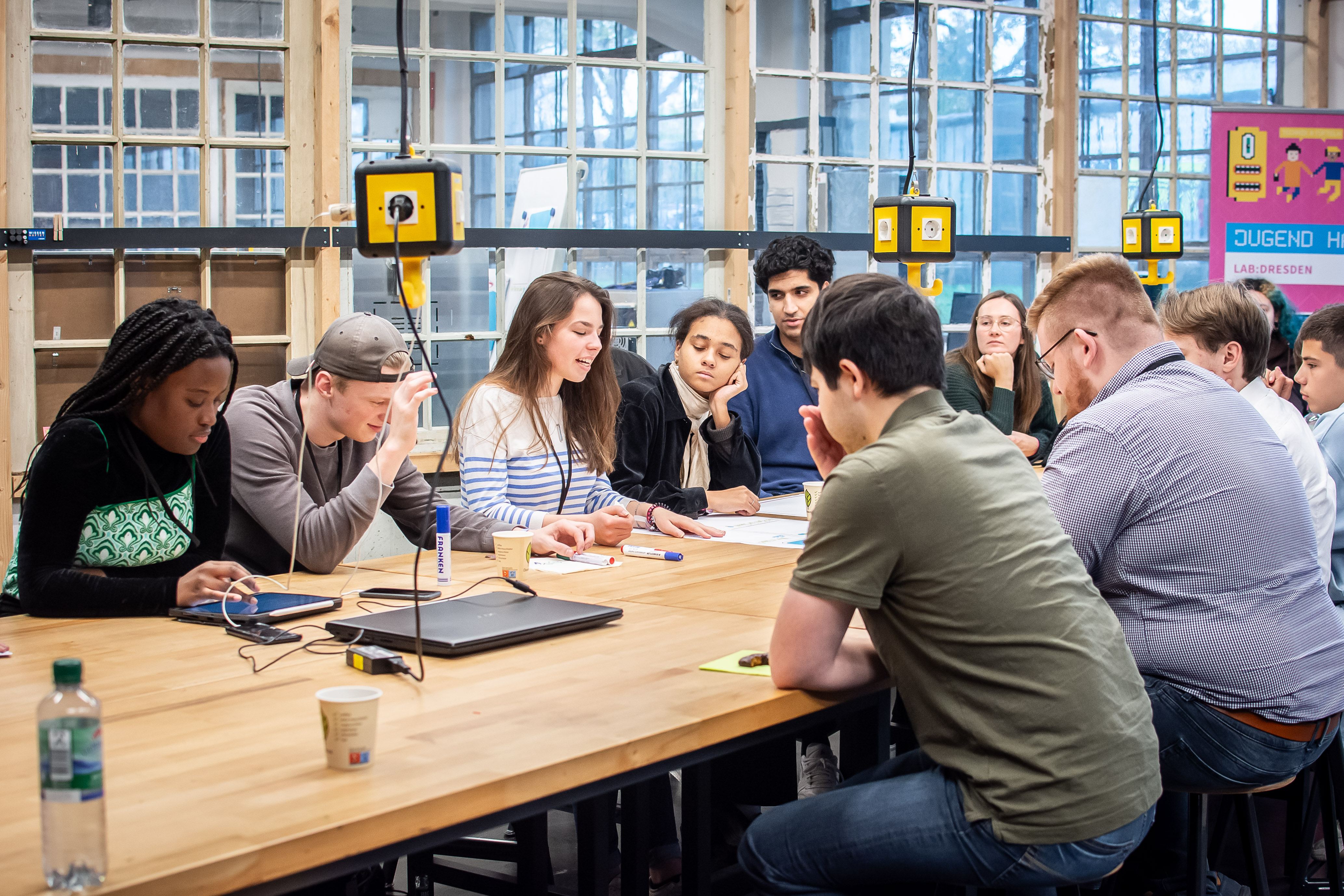 Auf dem Foto sieht man mehrere junge Menschen, die an einem großen Tisch in einem Büro sitzen und etwas besprechen. Auf dem Foto sieht man mehrere junge Menschen, die an einem großen Tisch in einem Büro sitzen und etwas besprechen.