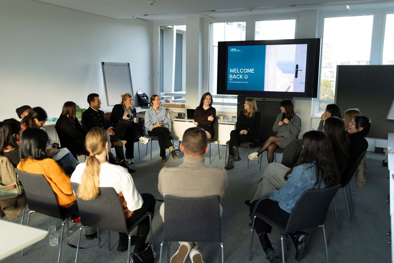 Menschen sitzen in einem hellen Seminarraum in einem Stuhlkreis und beteiligen sich an einer Besprechung. Auf einem Bildschirm steht „Welcome Back“. Das Bild zeigt eine einladende Atmosphäre für Gruppenarbeit oder Workshops.