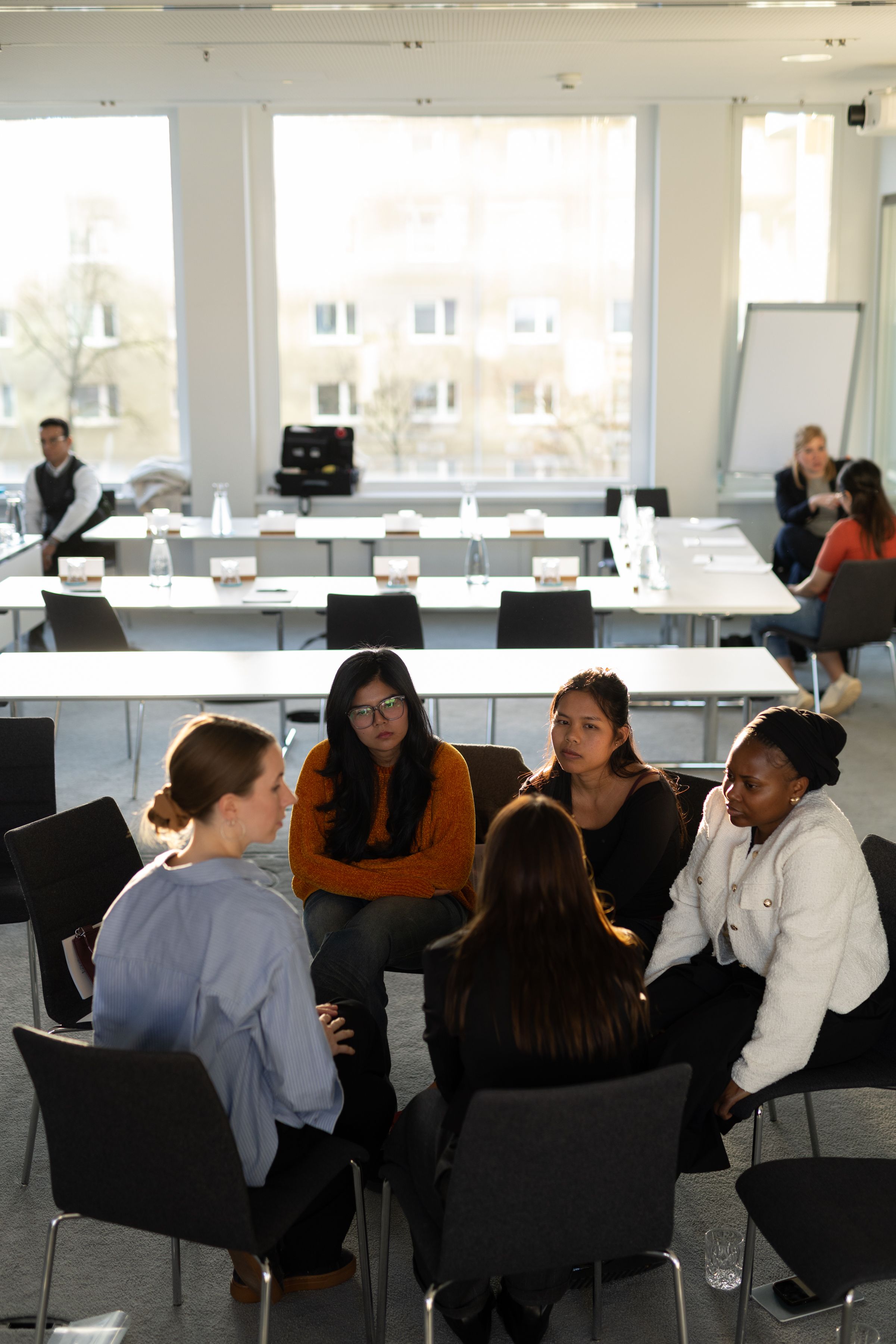 Fünf Personen sitzen in einem hellen Besprechungsraum im Stuhlkreis und führen eine konzentrierte Gruppendiskussion. Im Hintergrund sind leere Tische und weitere Teilnehmende des Meetings sichtbar.