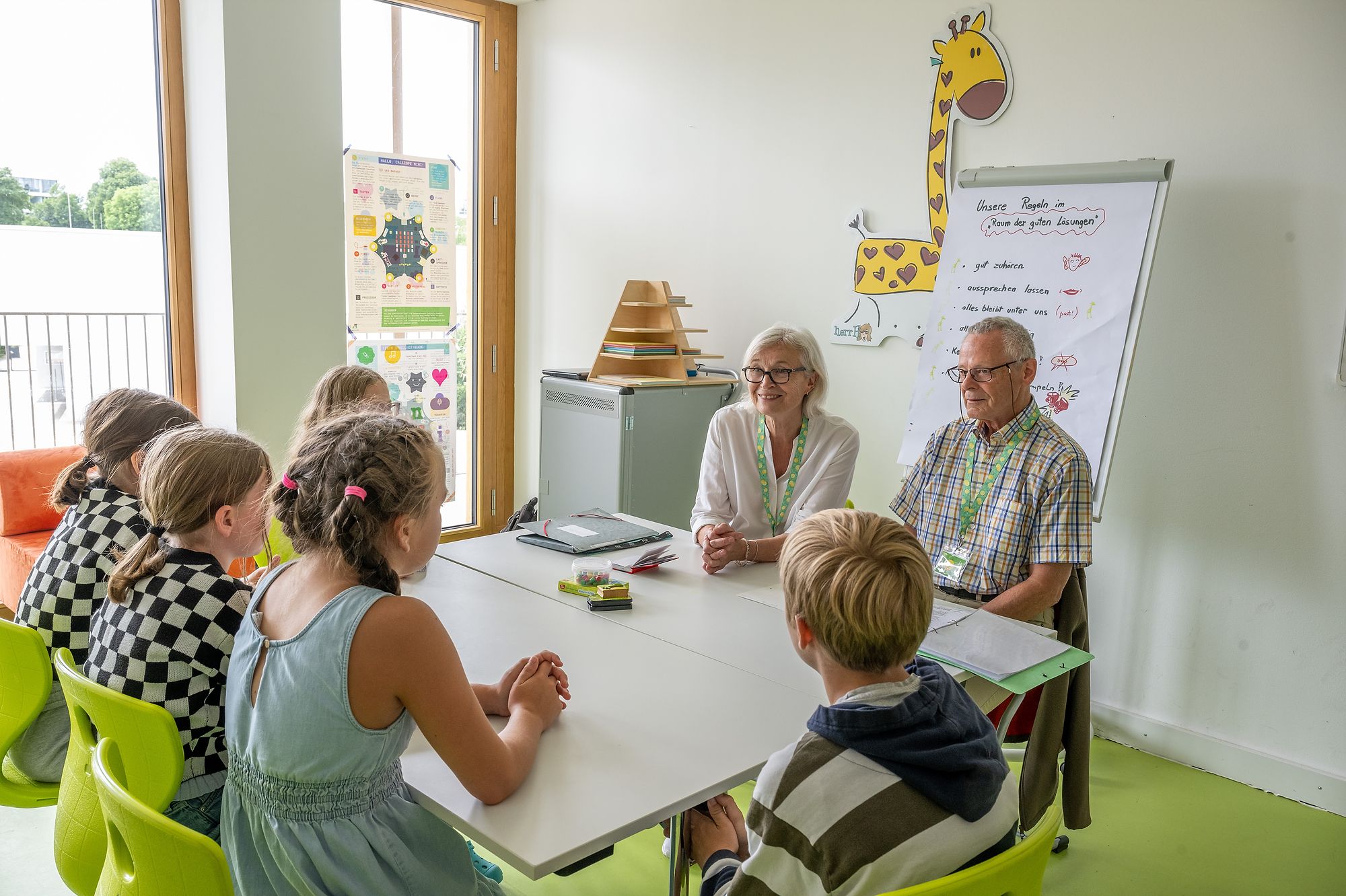Auf dem Foto sieht man vier Kinder, die in einem Klassenzimmer mit zwei älteren Menschen reden.