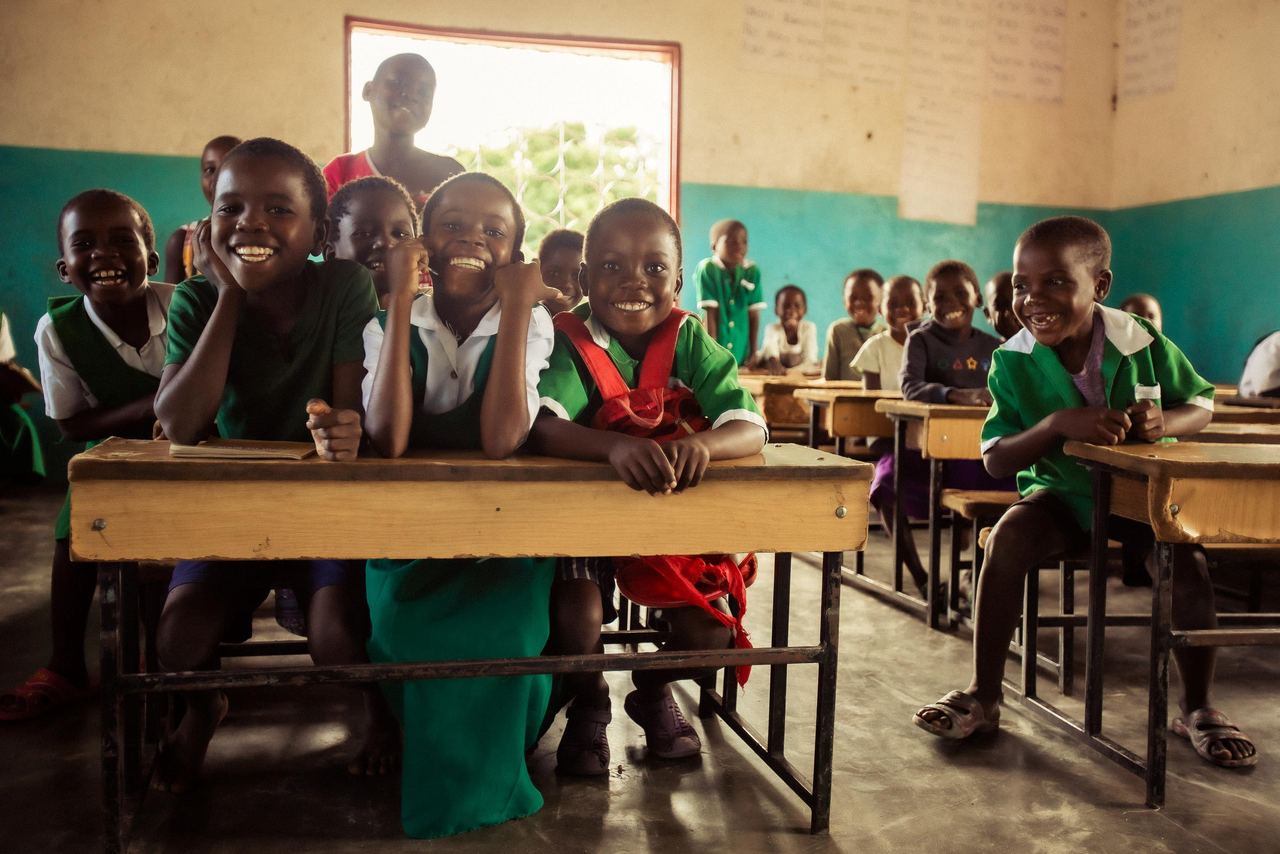 Fröhliche Schulkindern sitzen in einer hellen Klasse, lachen und blicken erwartungsvoll zur Kamera, symbolisieren Bildung und Gemeinschaft.