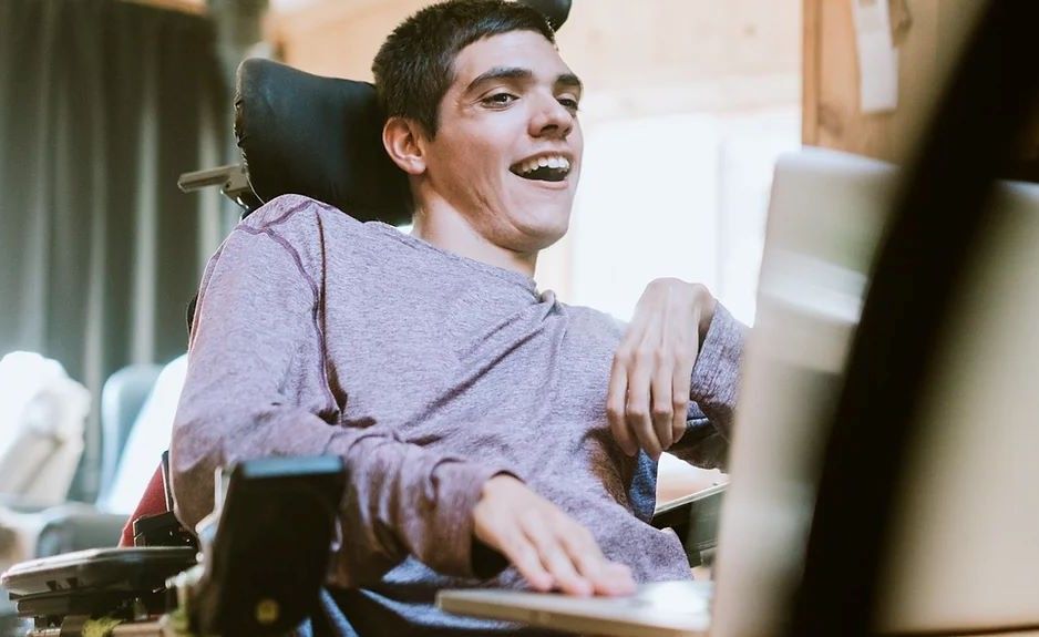 Junger Mann im Rollstuhl arbeitet entspannt am Laptop in einem gemütlichen, hellen Wohnraum.