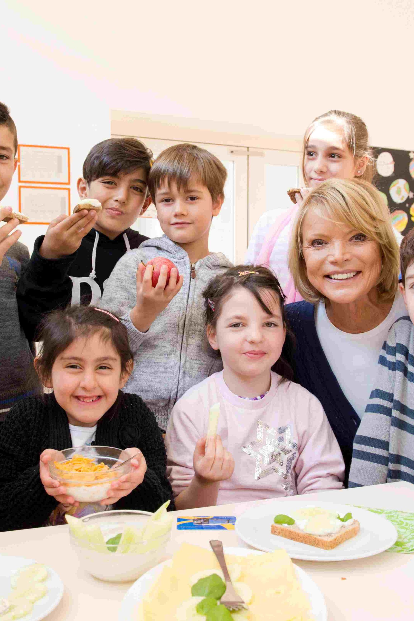 Eine fröhliche Gruppe von Kindern und einer erwachsenen Frau sitzt gemeinsam an einem Tisch und genießt gesunde Snacks in einer hellen, freundlichen Umgebung.