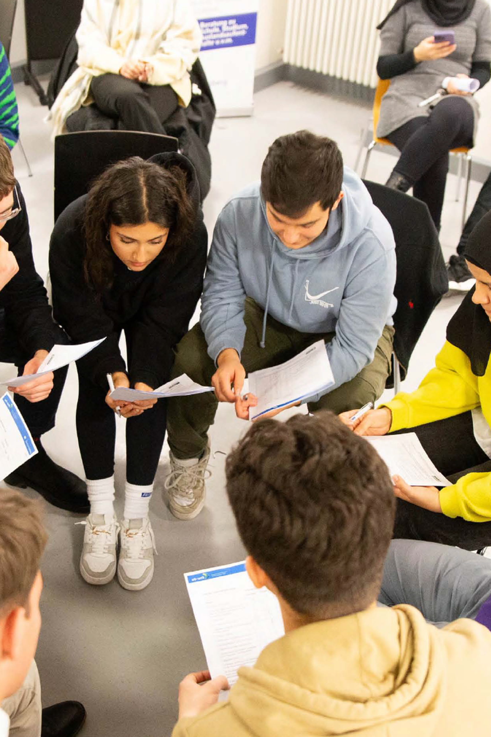 Das Foto zeigt junge Menschen, die in einem Stuhlkreis zusammensitzen und auf Blätter in ihren Händen gucken.
