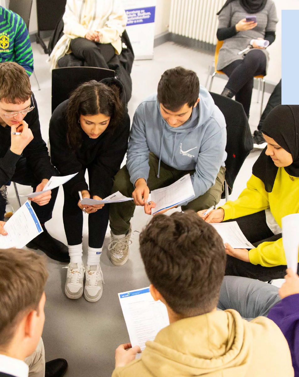 Das Foto zeigt junge Menschen, die in einem Stuhlkreis zusammensitzen und auf Blätter in ihren Händen gucken. Das Foto zeigt junge Menschen, die in einem Stuhlkreis zusammensitzen und auf Blätter in ihren Händen gucken.