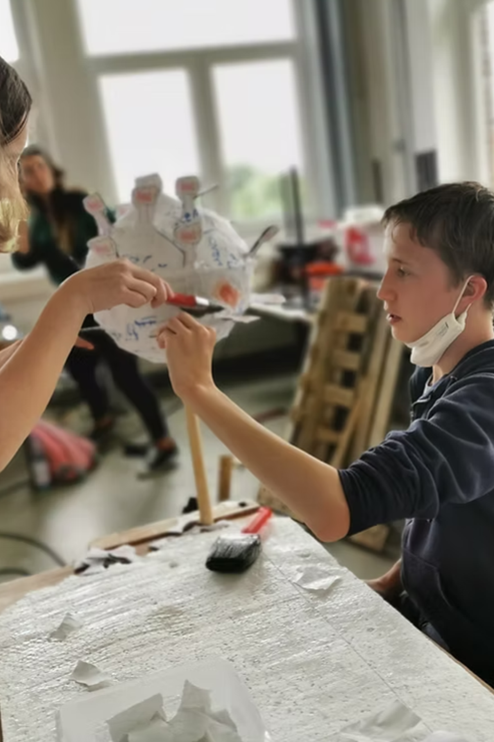 Das Foto zeigt einen jungen Teenager im Kunstunterricht, der etwas von einer jungen Frau gereicht bekommt.