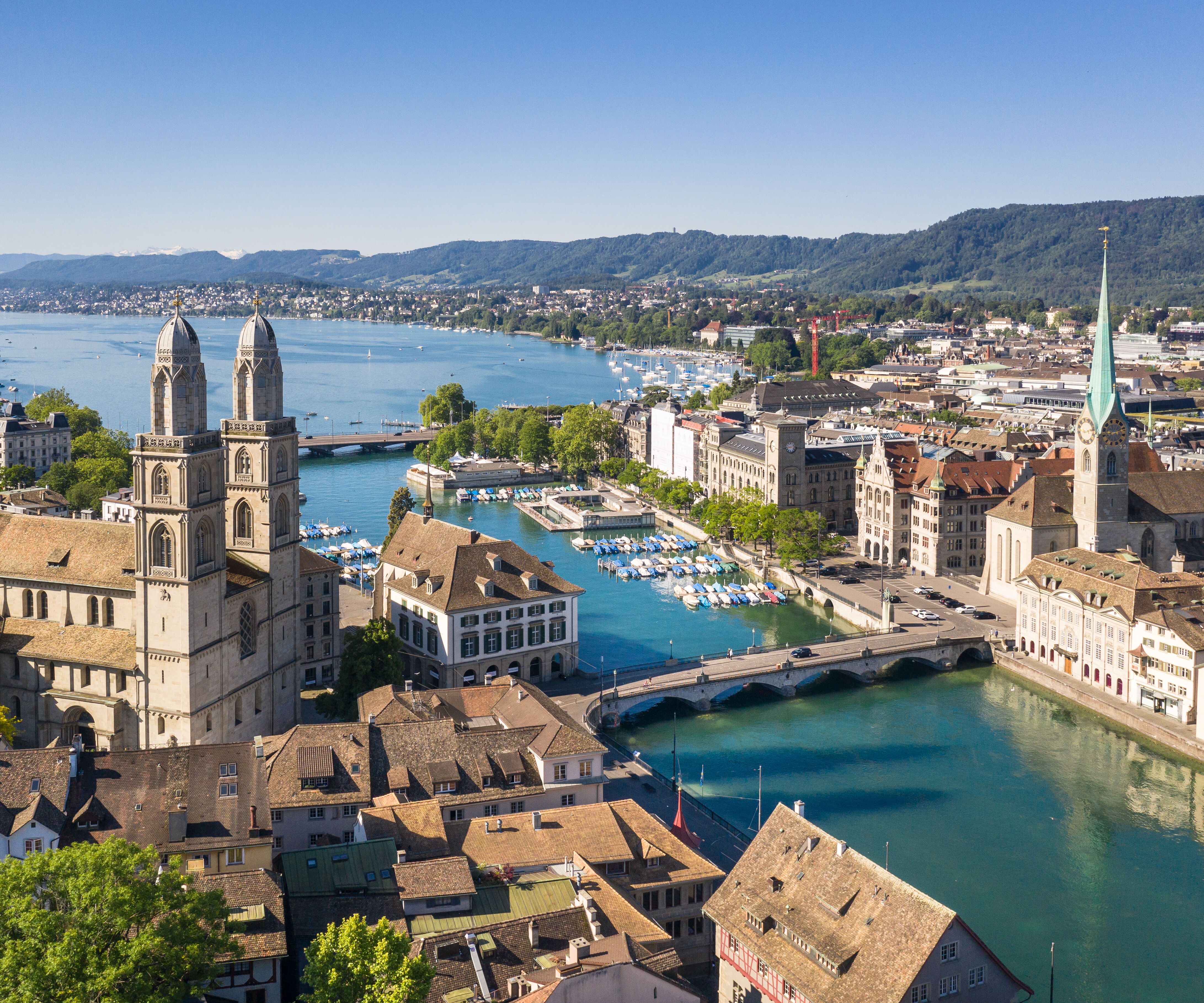 Blick auf die Altstadt Zürichs mit der Limmat, historischen Gebäuden, der Kirche Großmünster und dem Flussufer, eingebettet zwischen Seen und Hügeln bei klarem Himmel.