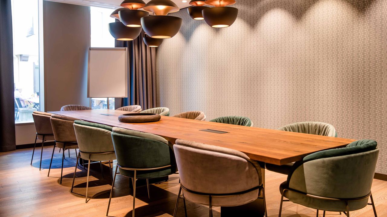 Motel One Konferenzraum mit großem Holztisch, komfortablen Stühlen und stilvollen Hängeleuchten.