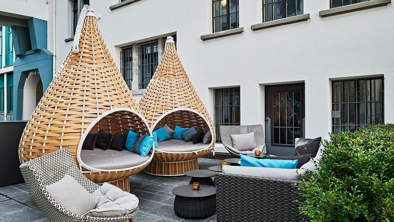 Gemütliche Außenlounge mit zwei großen, hängenden Korbsesseln, Polstern in Blau- und Grautönen, sowie Sofas und einem kleinen Tisch. Atmosphäre für entspannte Momente im Innenhof eines Gebäudes.