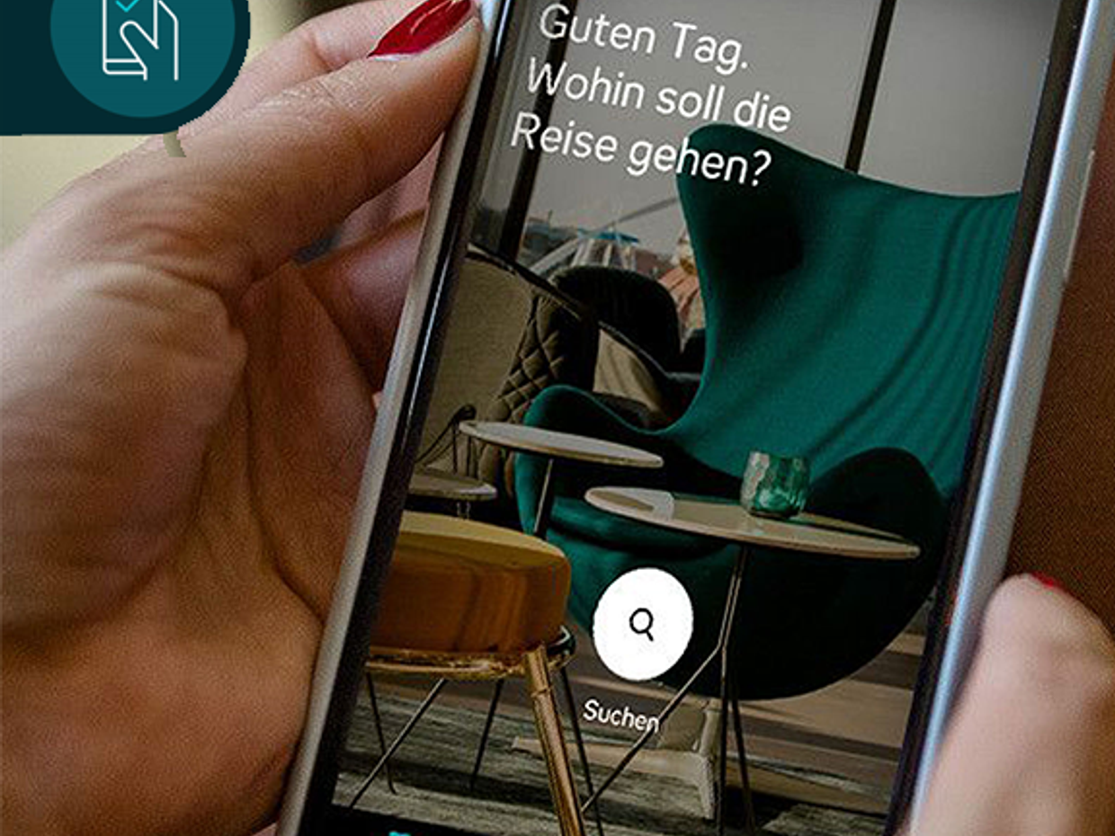 Person hält Smartphone, auf dem eine Reisebuchungs-App mit der Frage "Guten Tag. Wohin soll die Reise gehen?" und einer Suchfunktion zu sehen ist.