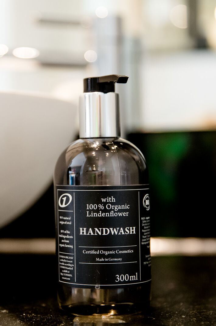 Flasche mit flüssiger Handseife aus 100 % biologischer Lindenblüte neben weißem Waschbecken auf schwarzer Oberfläche, modernes Badezimmerambiente. Flasche mit flüssiger Handseife aus 100 % biologischer Lindenblüte neben weißem Waschbecken auf schwarzer Oberfläche, modernes Badezimmerambiente.