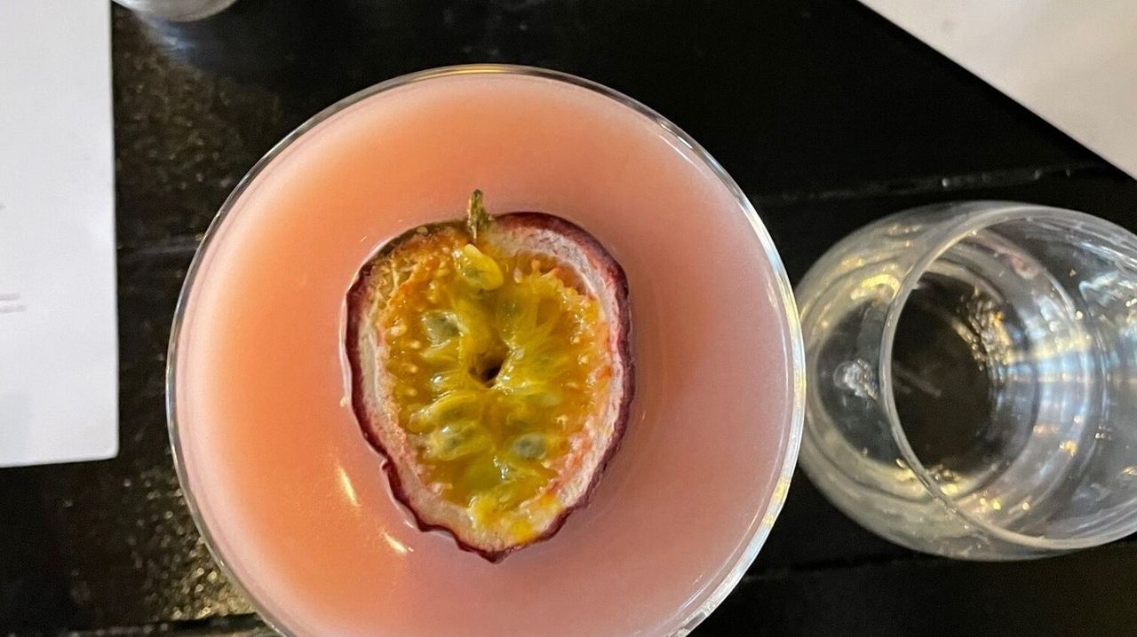 Ein rosa Cocktail in einem Glas, garniert mit einer halbierten Passionsfrucht, daneben Salz- und Pfefferstreuer sowie ein Wasserglas und eine Speisekarte auf einem dunklen Tisch.