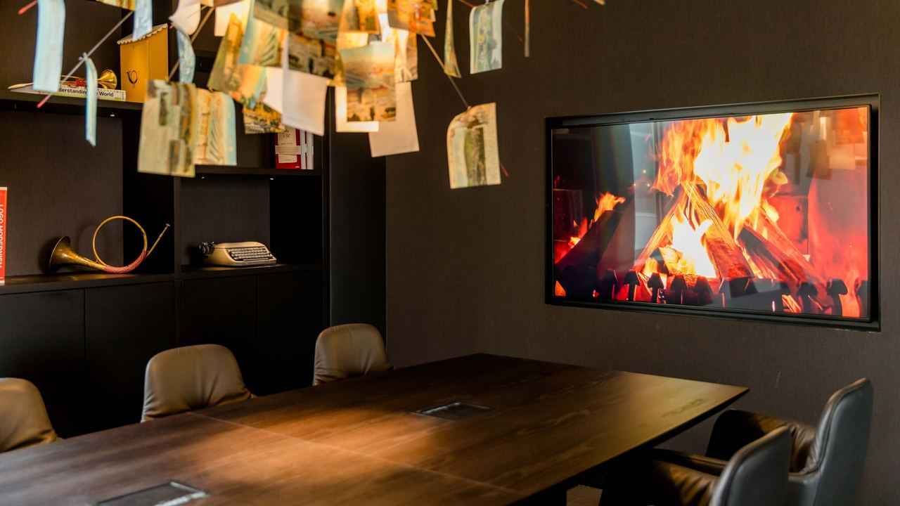 Motel One Besprechungsraum mit großem Holztisch, bequemen Stühlen und einem modernen Kaminfeuer an der Wand. Über dem Tisch hängt eine kreative, kunstvolle Deckenlampe aus vielen Papierblättern.