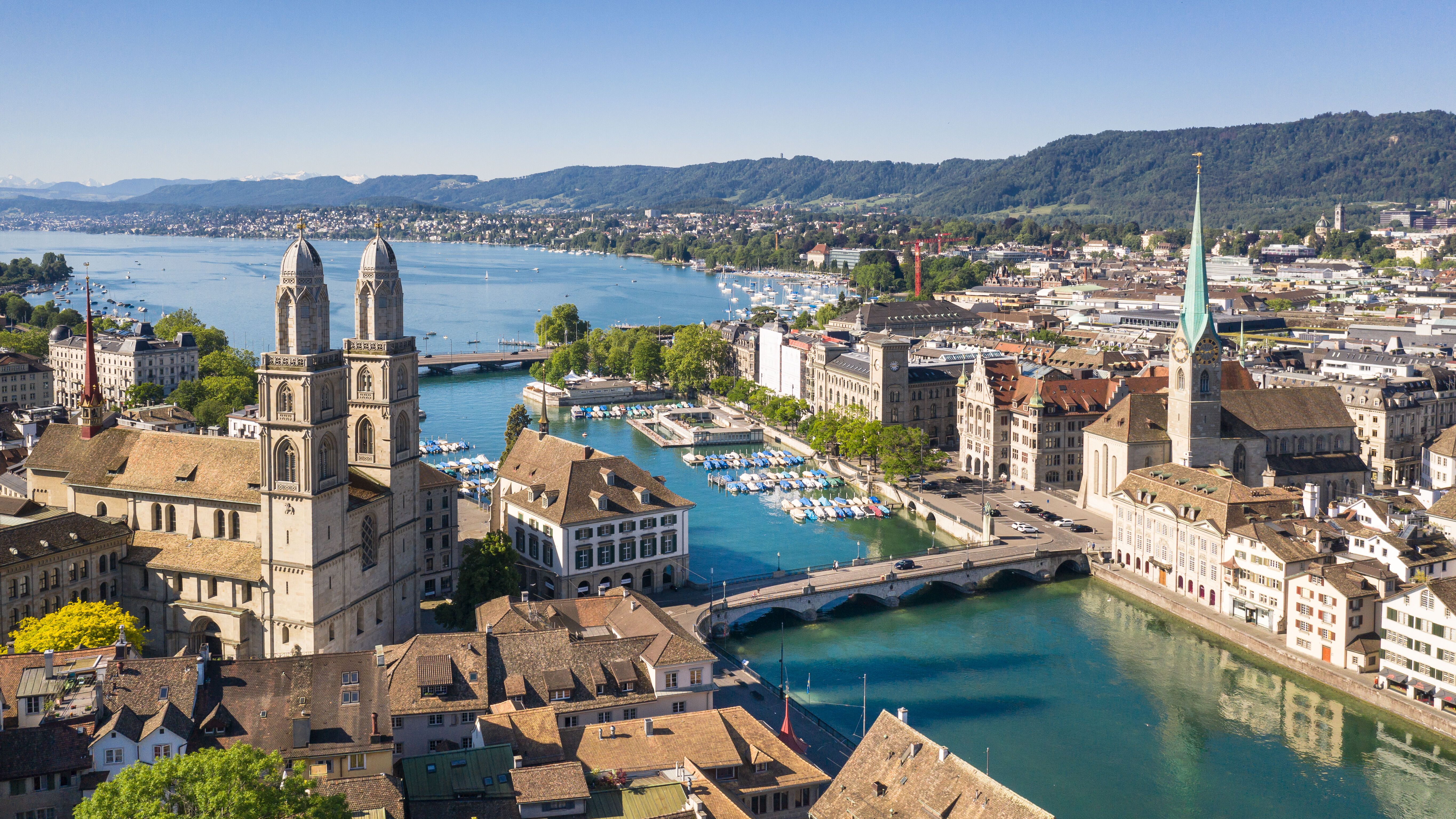 Panoramablick auf Zürich mit historischem Stadtkern, Flusslauf, Brücken und Alpen im Hintergrund, bei klarem Himmel und sonnigem Wetter.